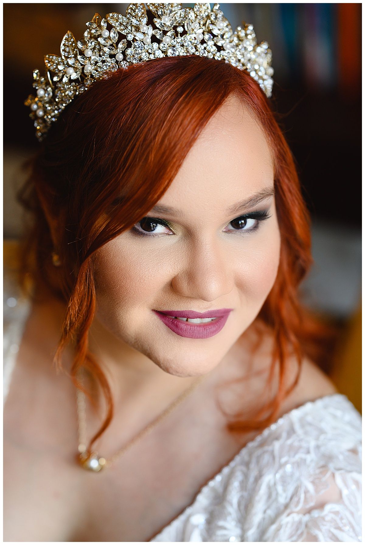 Red-haired bride wearing a crystal tiara and white lace dress with berry lips and elegant pearl necklace.