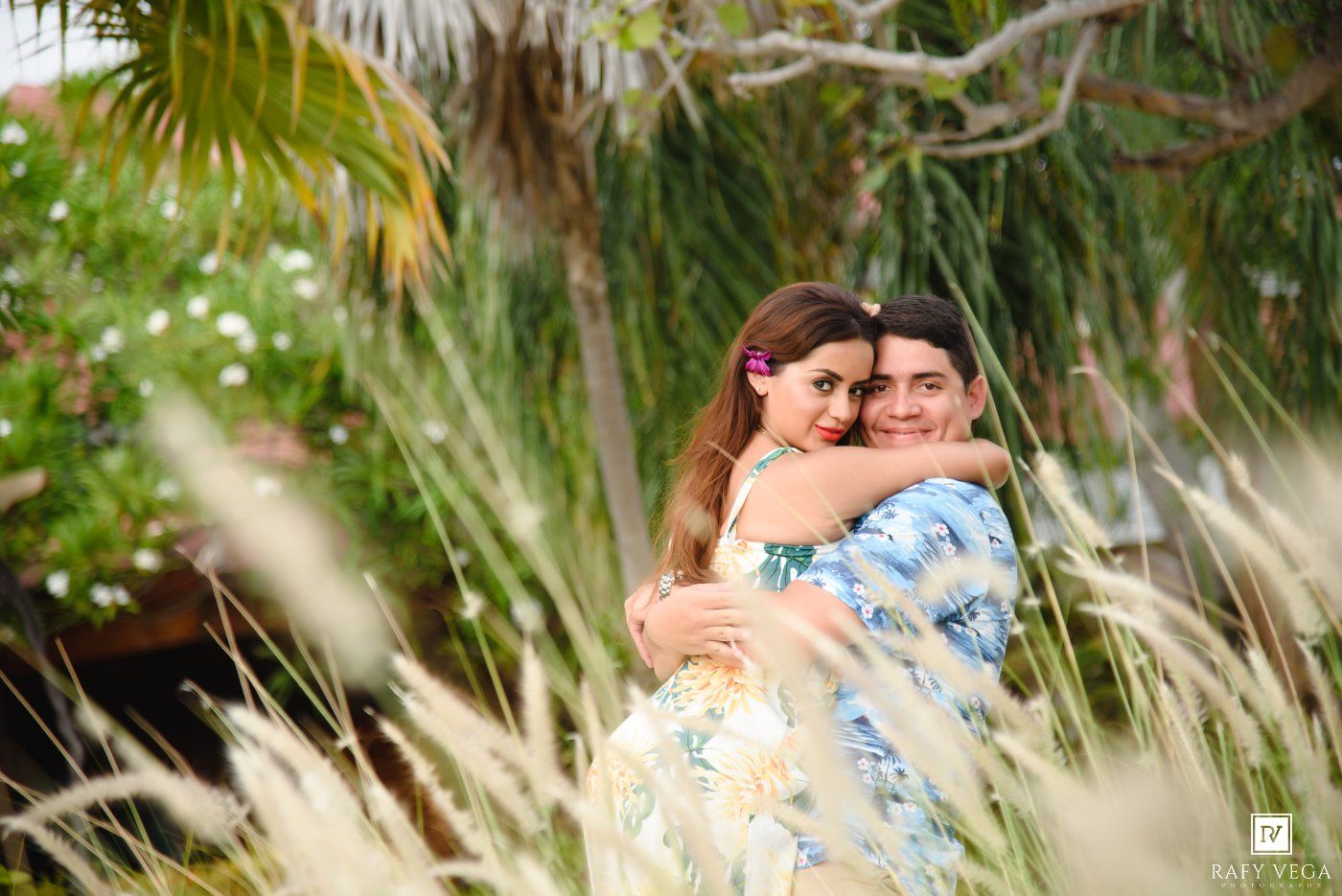 Two women share a warm embrace outdoors, surrounded by lush tropical greenery and tall ornamental grasses.