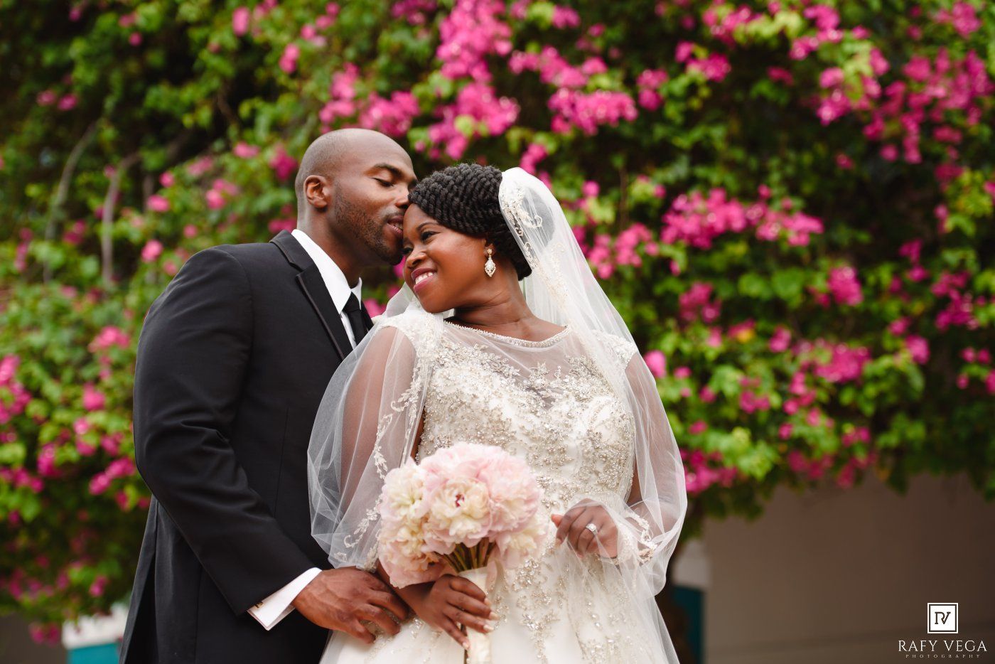Bride and groom share romantic moment before pink flowering bushes, bride in lace gown holding blush bouquet.