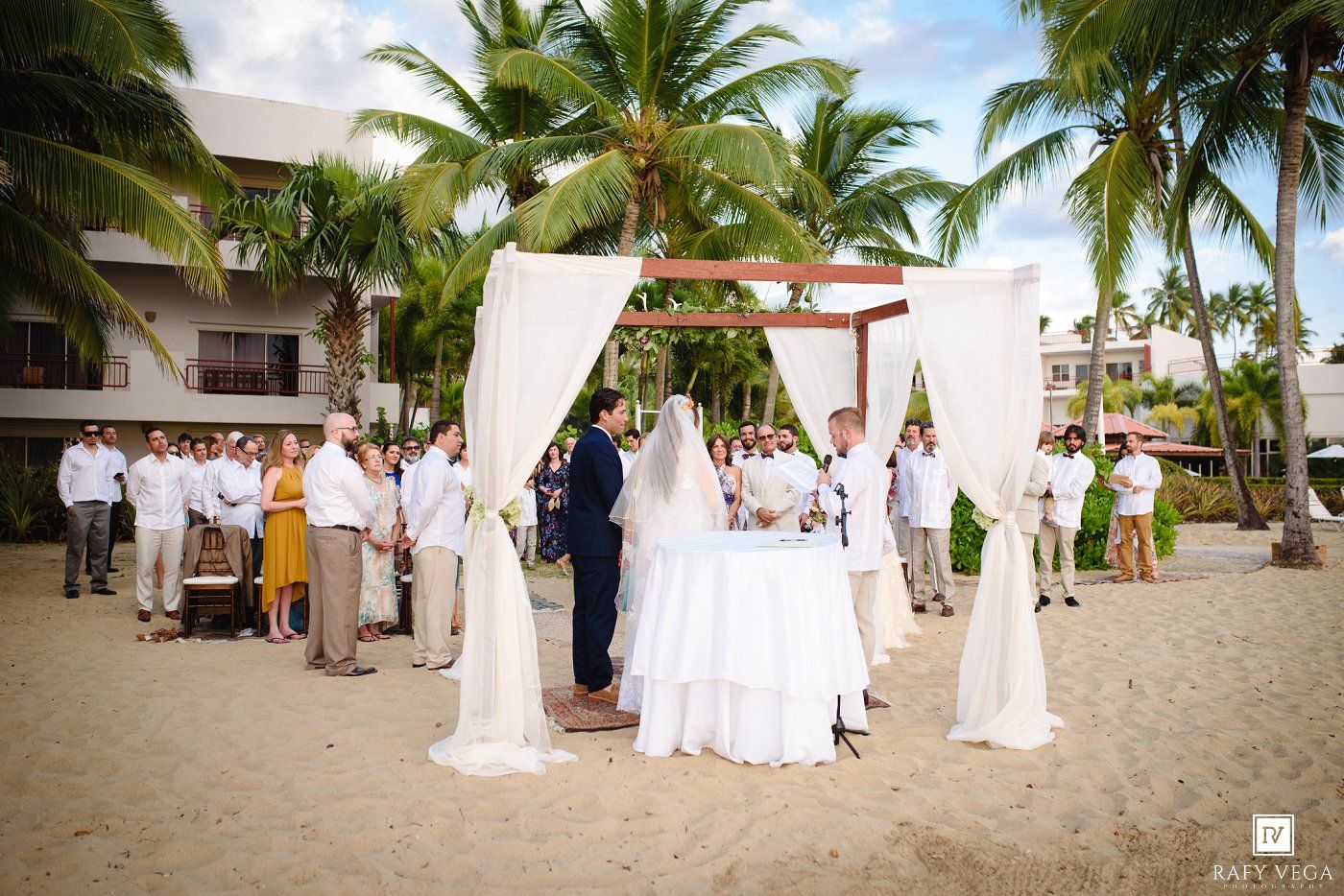Couple exchanges vows at elegant outdoor beach wedding ceremony under white draped canopy with palm trees.