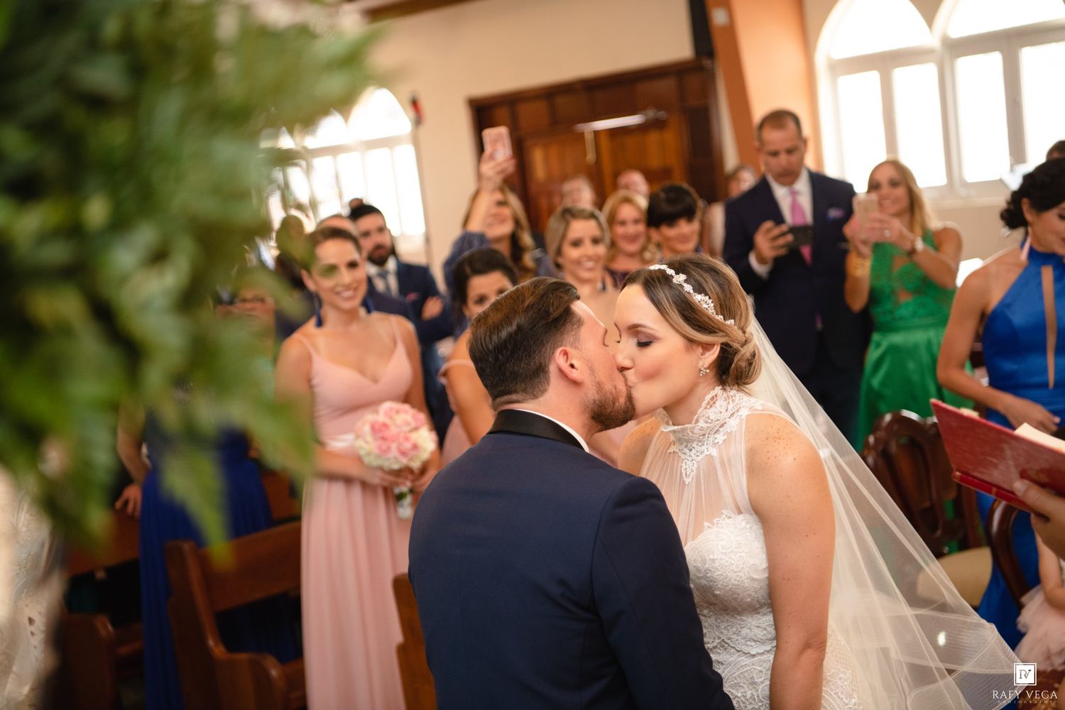 Bride and groom share first kiss at church altar surrounded by bridesmaids in pink and wedding guests.