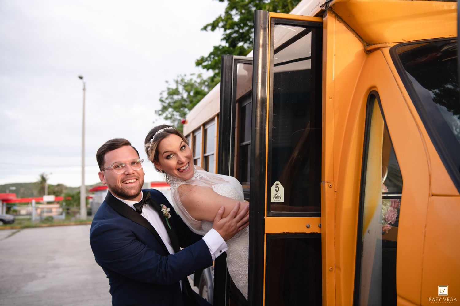 Bride and groom smiling together boarding a yellow school bus on their wedding day.