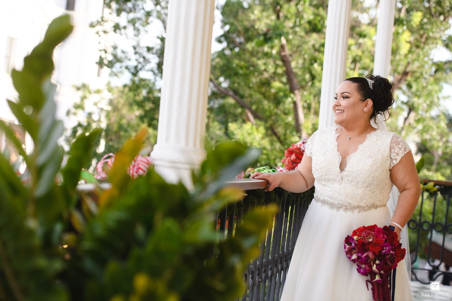 Bride in lace wedding dress holding red bouquet stands on elegant white columned veranda surrounded by tropical greenery.