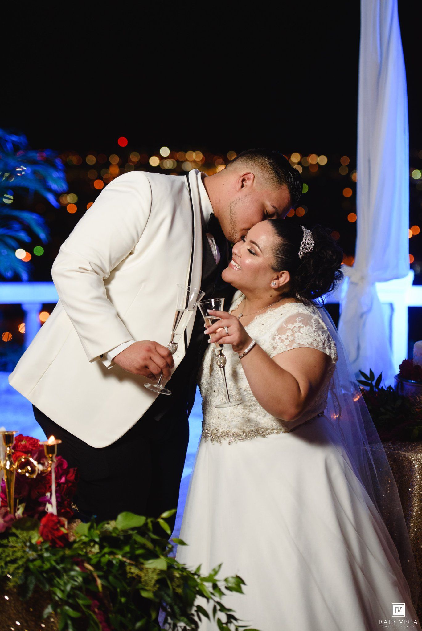 Bride and groom share a romantic kiss during their wedding reception toast at night with city lights in the background.