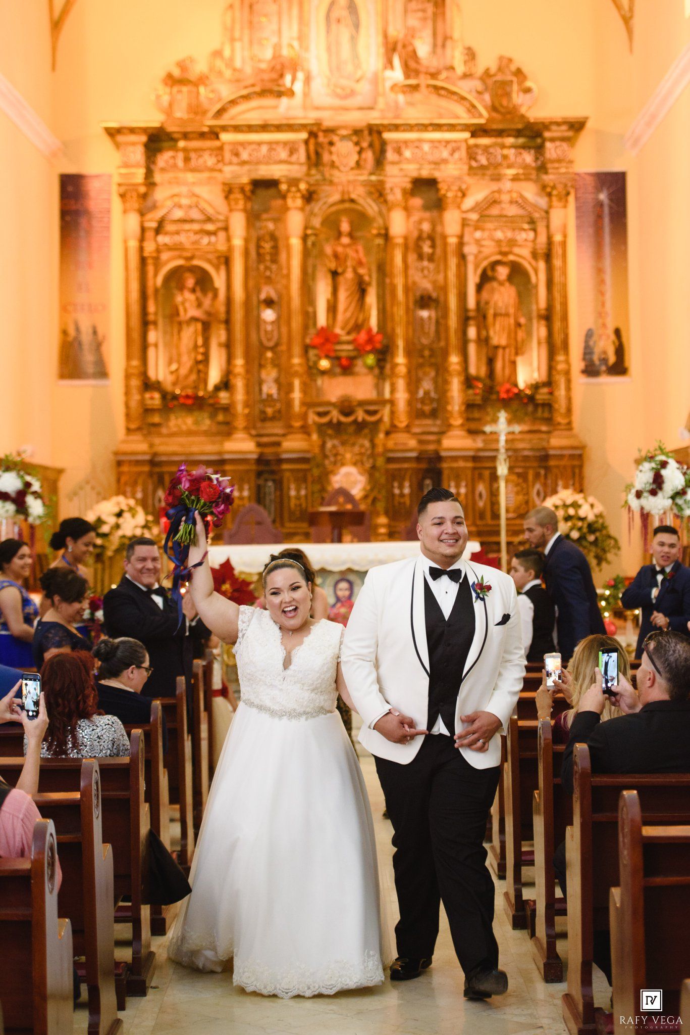 Joyful newlyweds walk down church aisle after ceremony, bride raising bouquet in celebration.