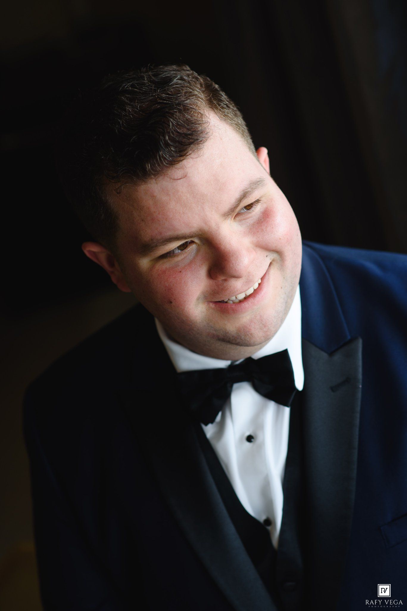 Smiling groom in navy blue tuxedo with black bow tie looking up at camera on wedding day.