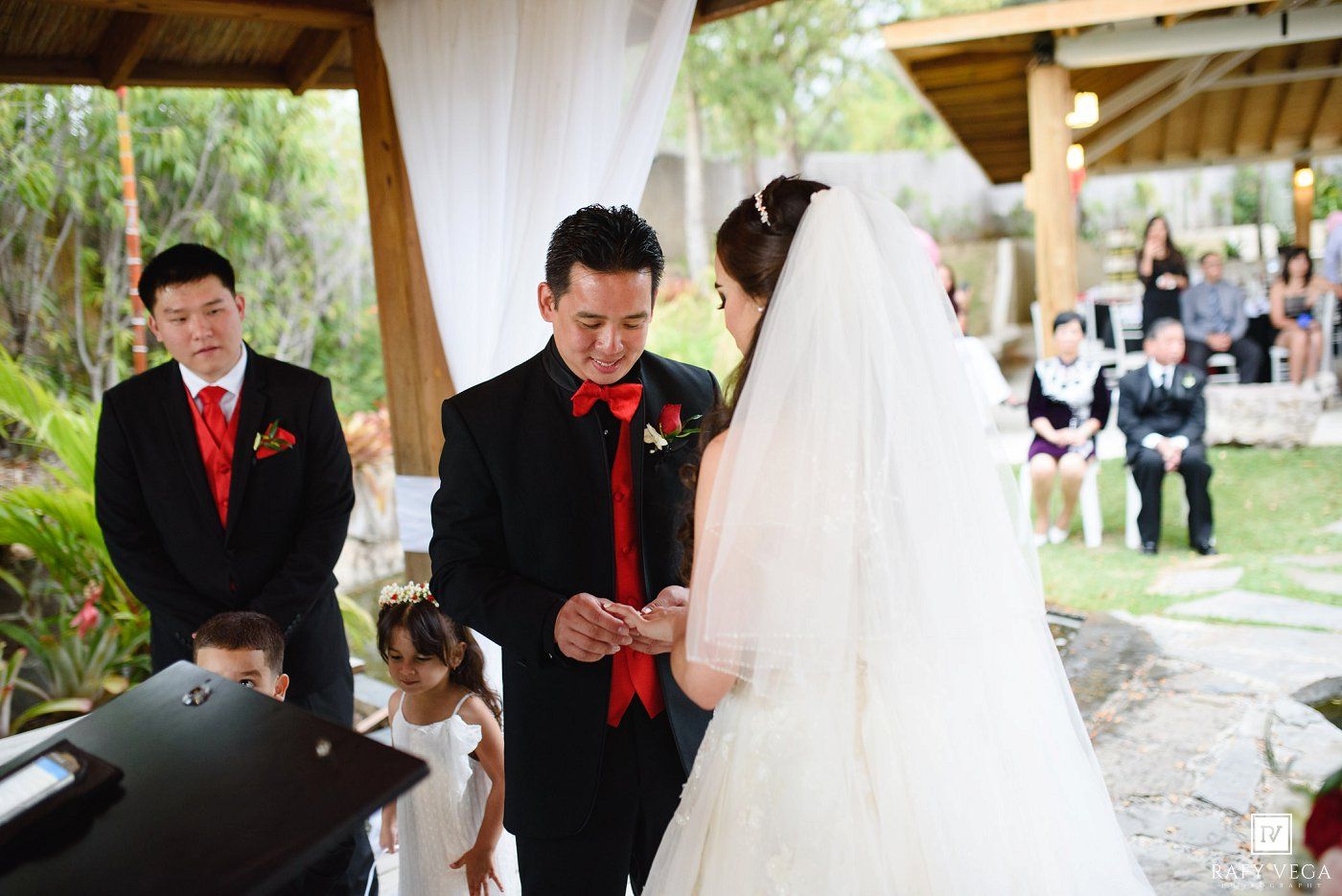 Asian couple exchanging rings at outdoor wedding ceremony, groom in black suit with red tie, bride in white gown.