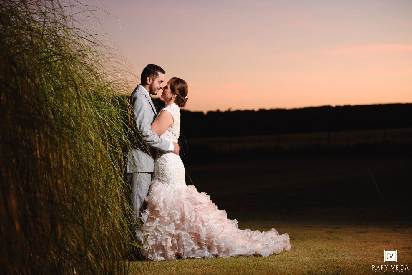 Bride and groom share romantic sunset embrace near tall grass in stunning outdoor wedding portrait.