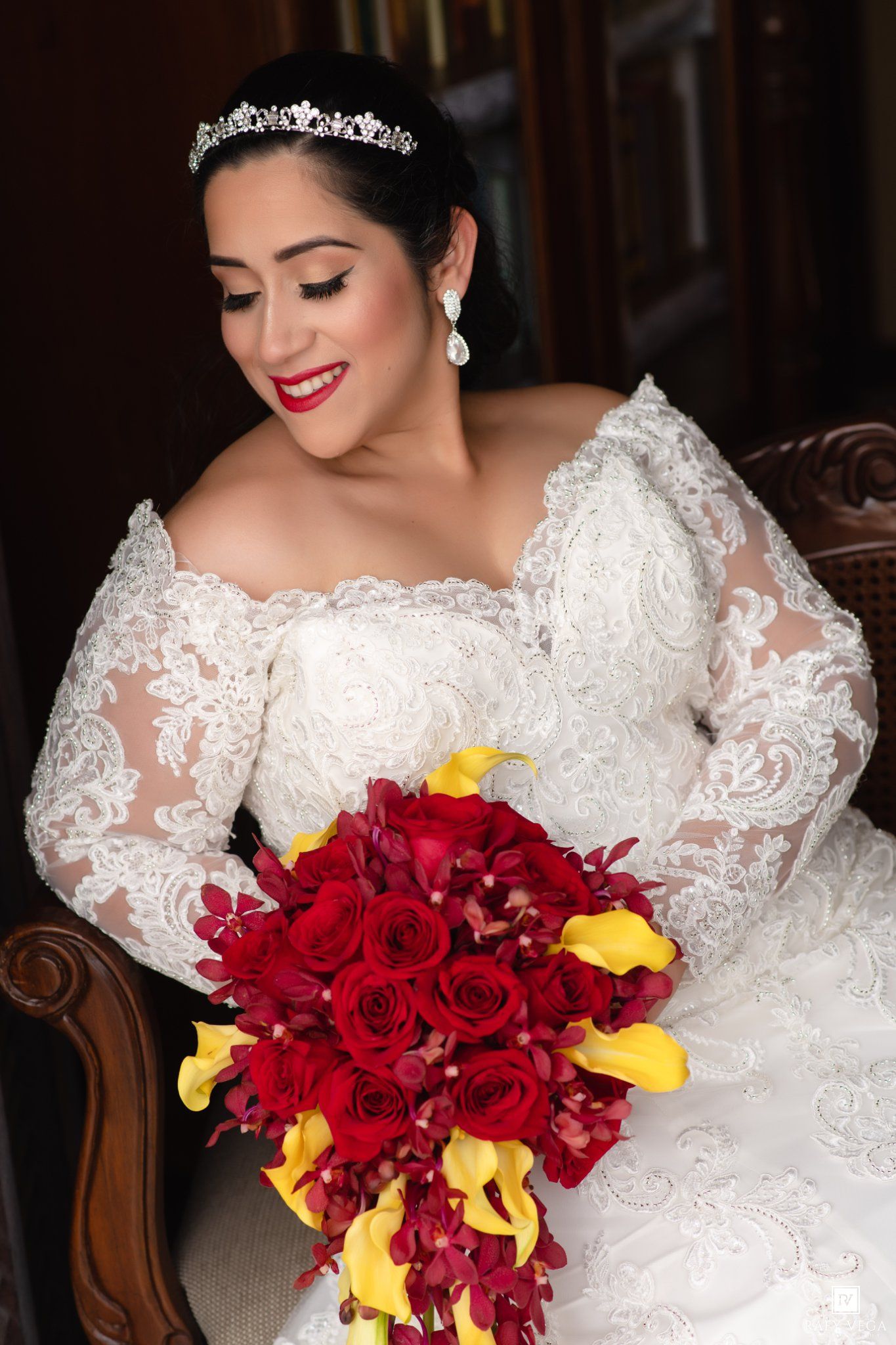 Bride in lace off-shoulder gown with tiara holds red rose and yellow calla lily bouquet while seated.