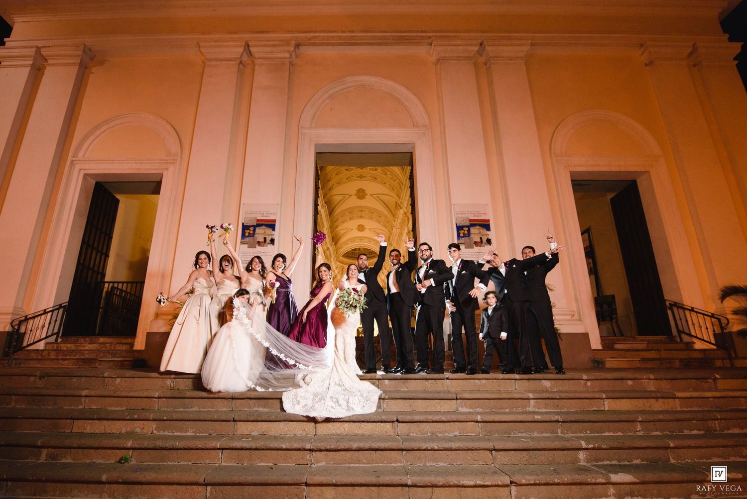 Wedding party celebrating on church steps at night, bride in white gown with bridesmaids and groomsmen posing joyfully.