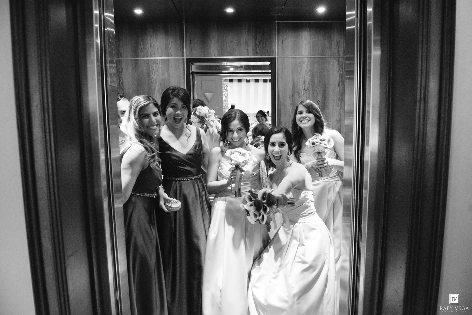 Black and white photo of a bride and bridesmaids smiling together inside an elevator on wedding day.