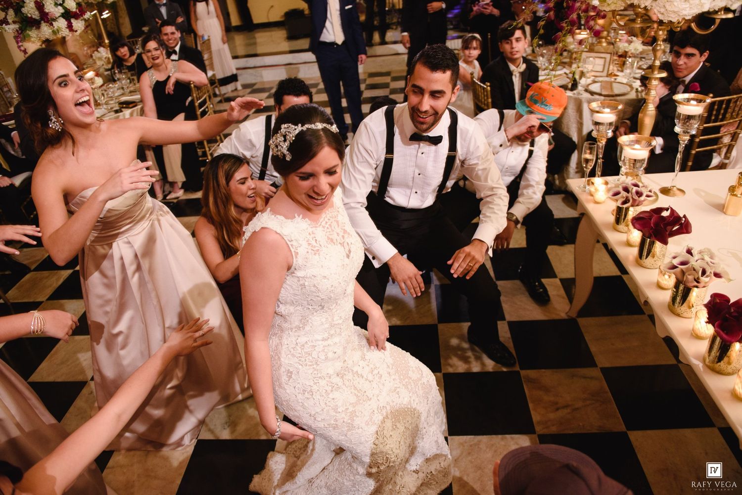 Bride and groom dancing joyfully with guests at elegant wedding reception on black and white checkered floor.