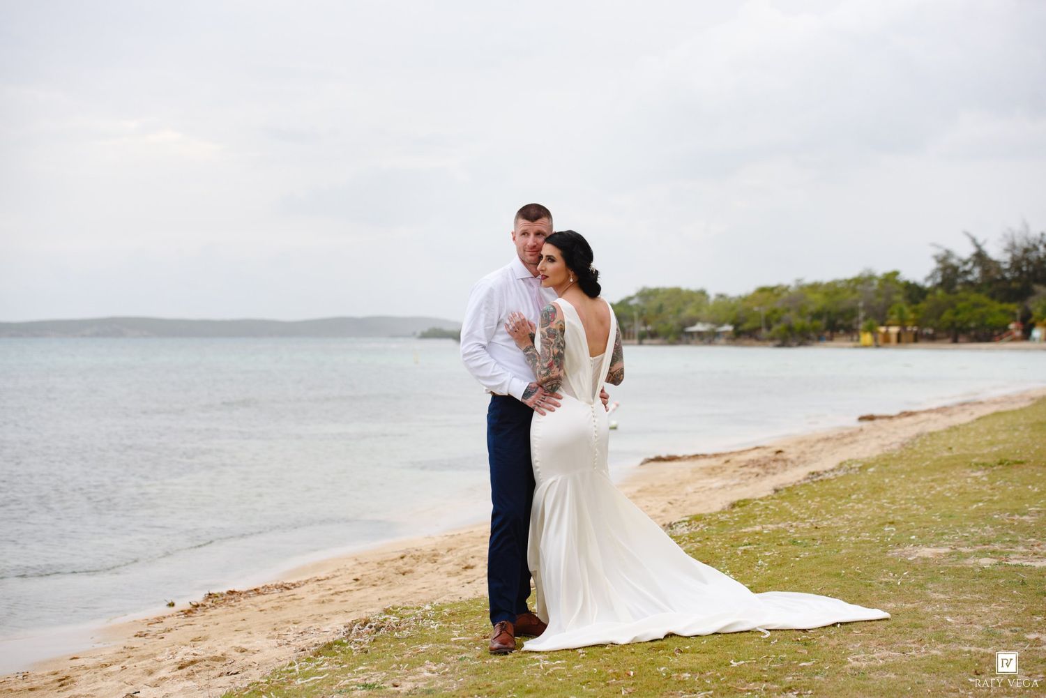 Groom embraces bride from behind on sandy beach, bride wearing fitted white mermaid gown with lace back detail.