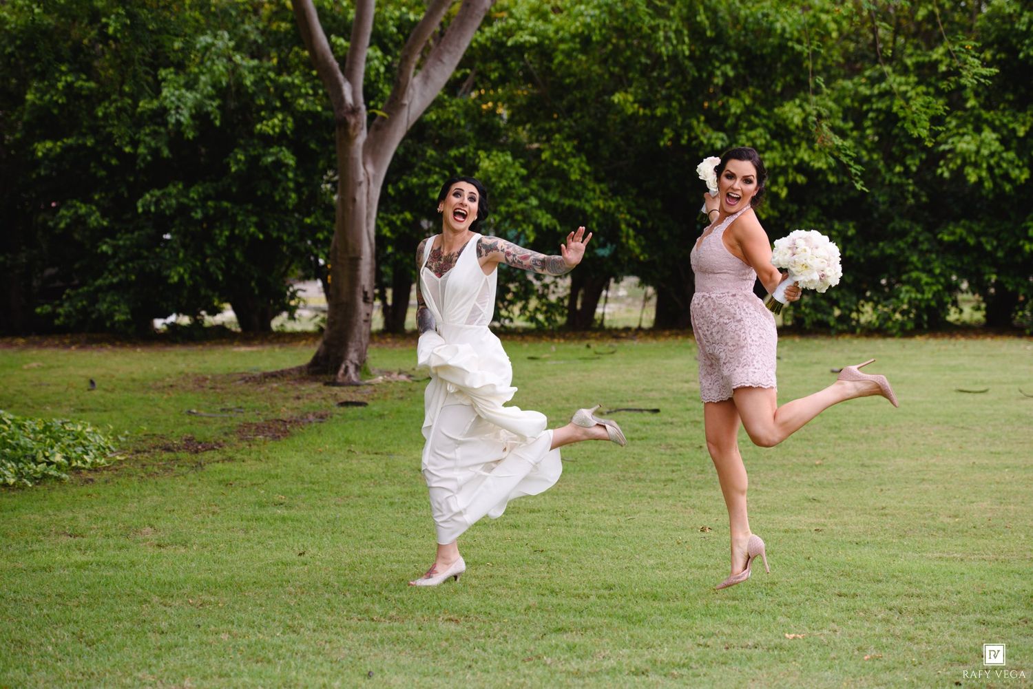 Bride and bridesmaid joyfully jumping on green lawn, bride in white gown with tattoos, bridesmaid holding white bouquet.