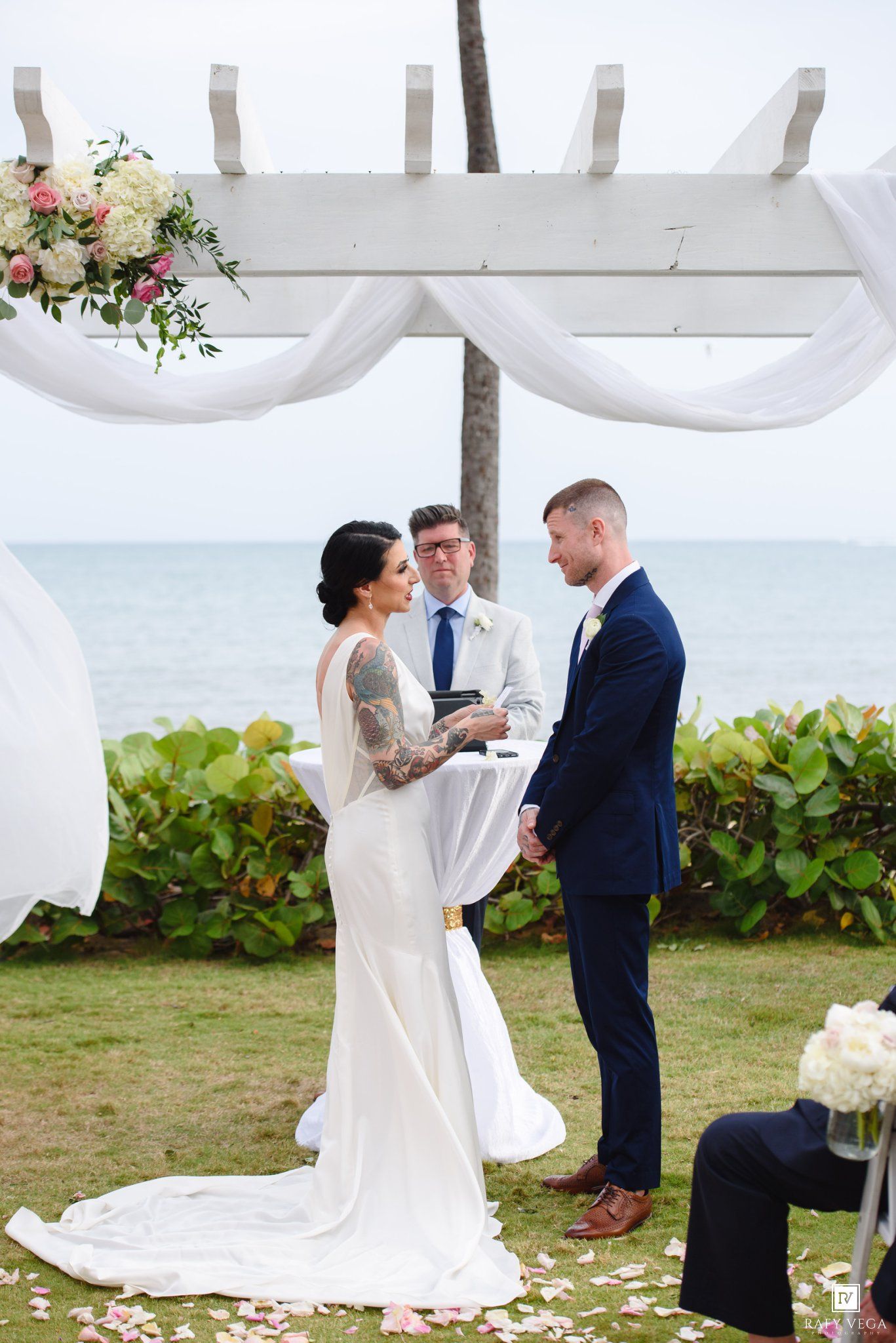 Bride and groom exchange vows at outdoor beach wedding ceremony under white floral pergola with ocean view.
