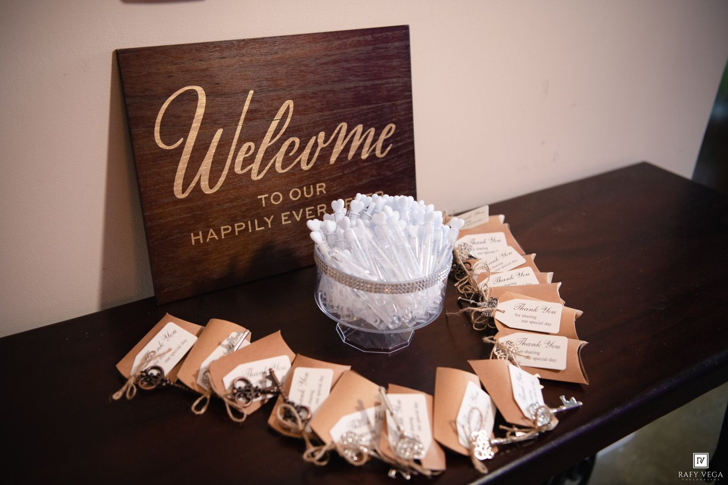 Wedding welcome table with kraft paper favor bags, white floral centerpiece, and rustic wooden Welcome to Our Happily Ever After sign.
