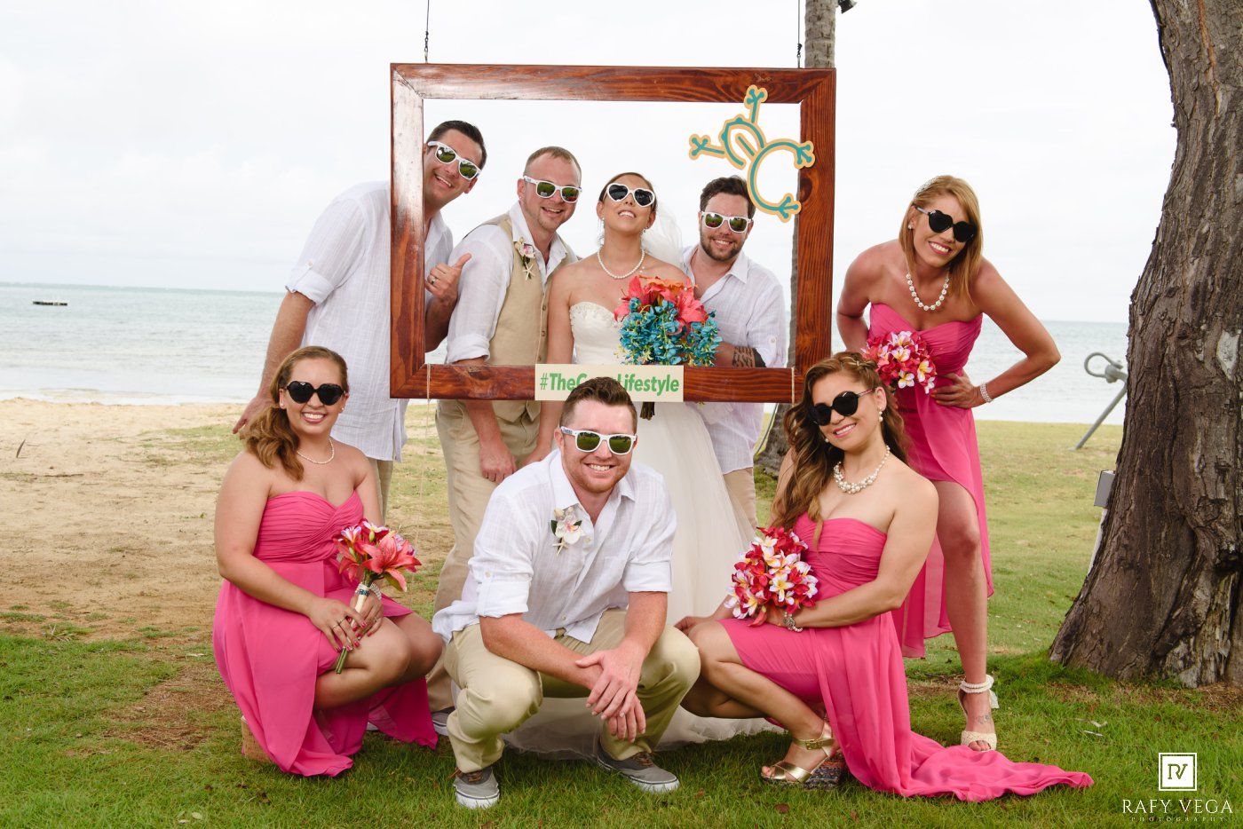 Wedding party posing in pink dresses with bouquets inside a wooden frame photo prop on a tropical beach.