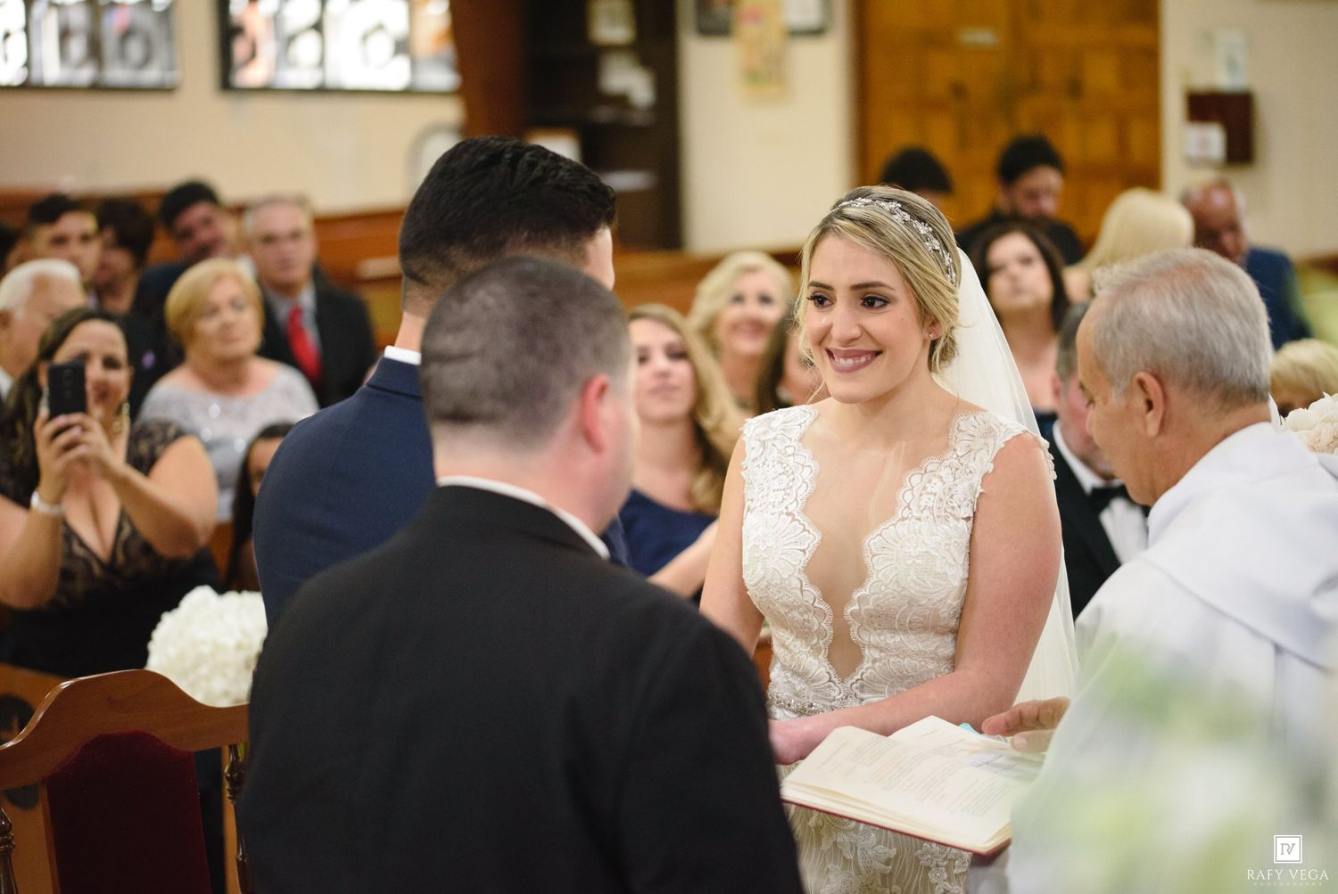 Smiling bride in lace gown exchanges vows with groom before guests at a church wedding ceremony.