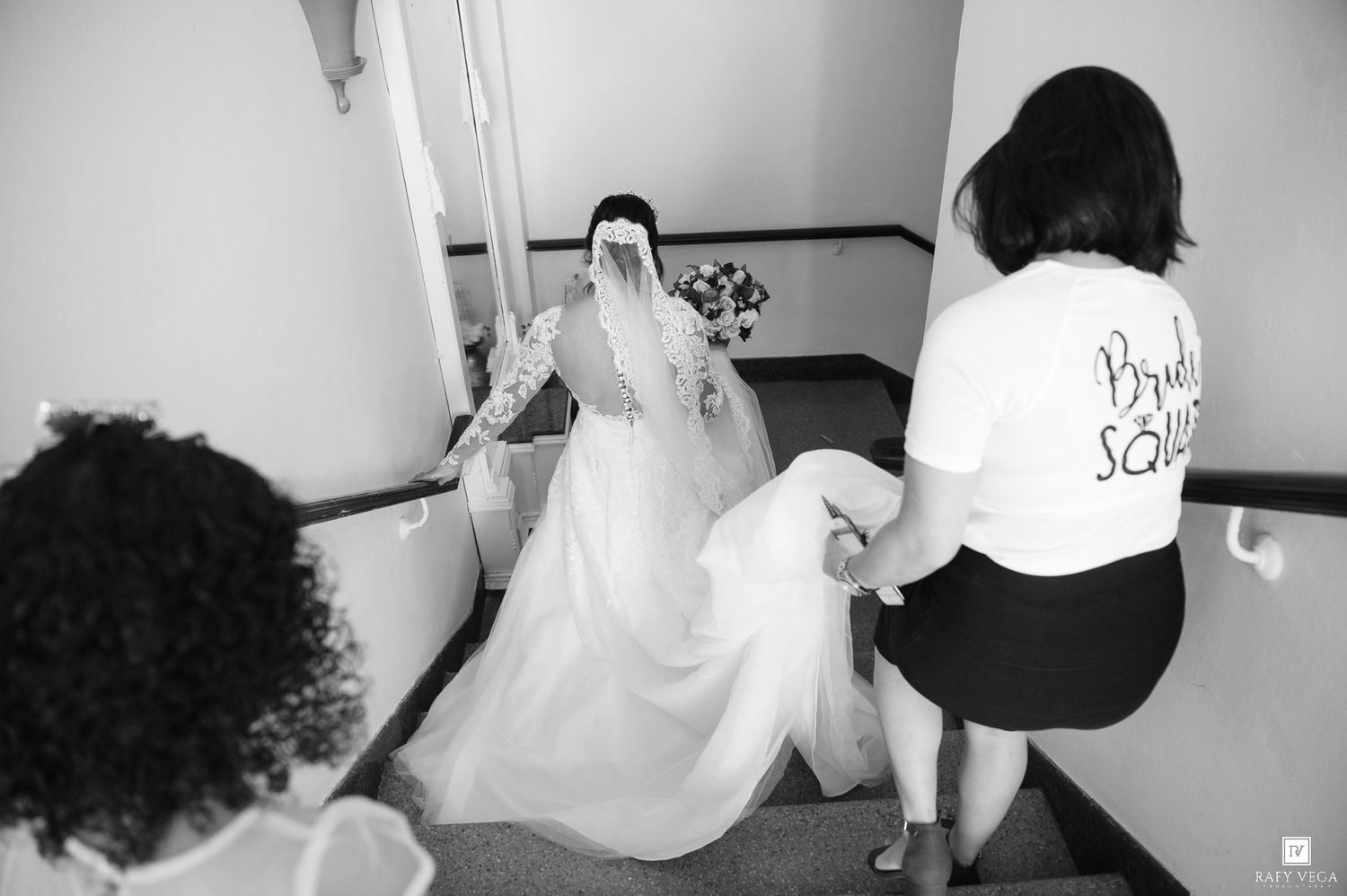 Bride in white gown descends stairs assisted by bridesmaids in black and white photo.