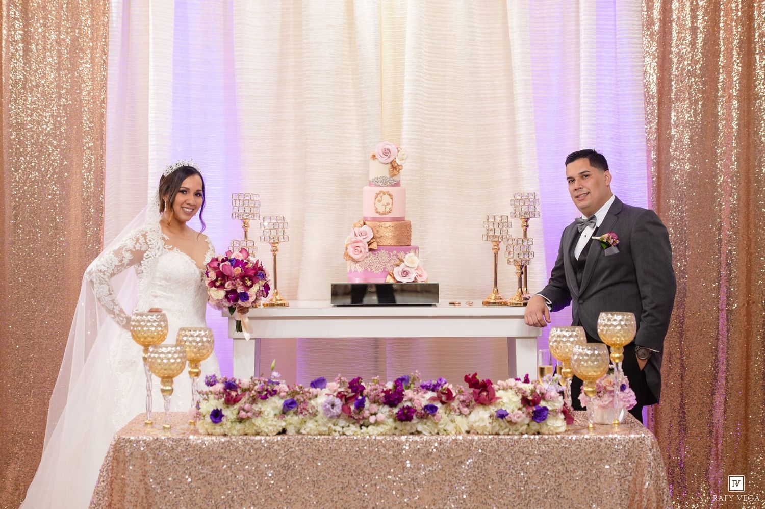 Bride and groom stand beside elegant wedding cake table with gold sequin tablecloth and purple floral arrangements.