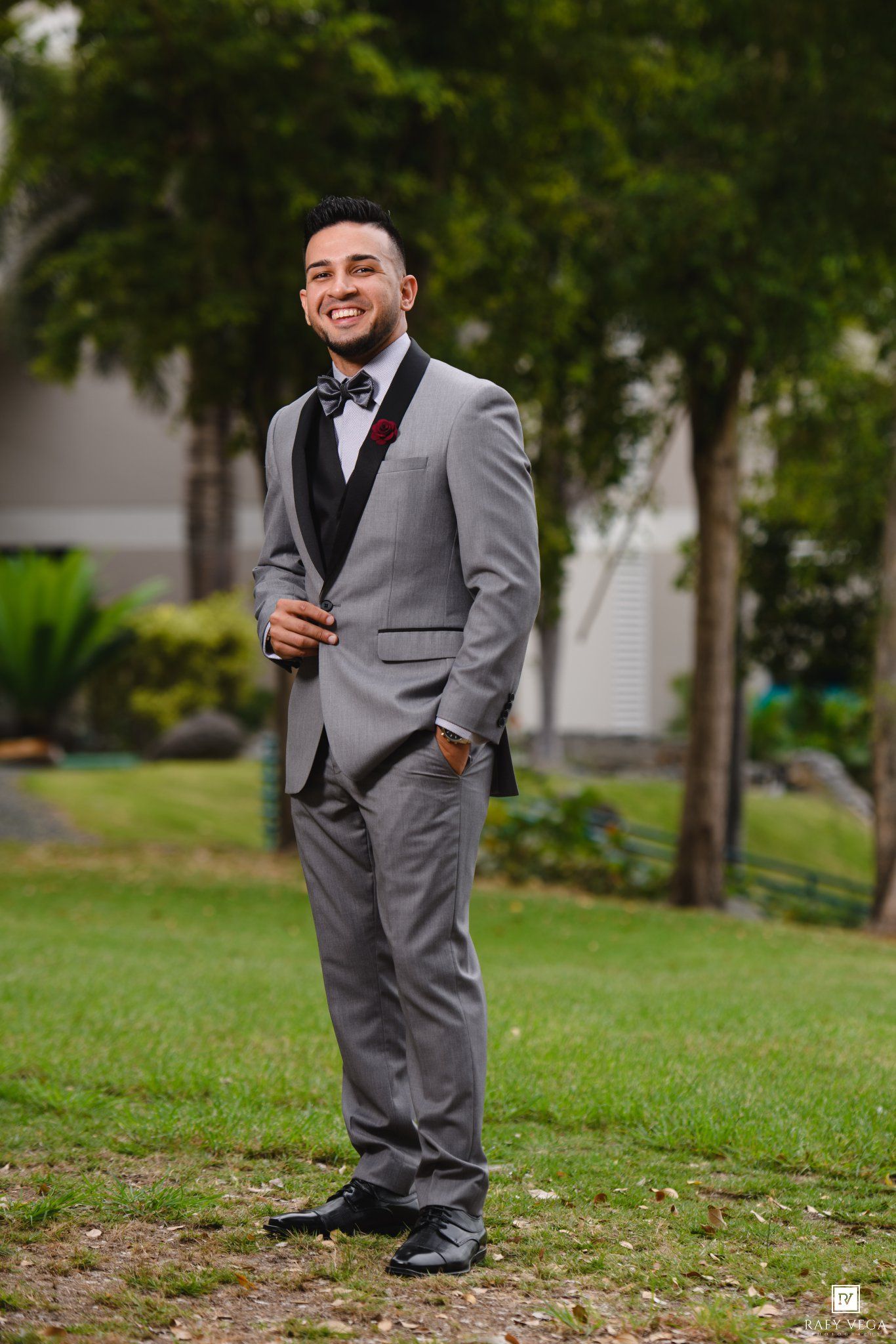 Smiling groom in gray suit with black vest and bow tie standing outdoors on green grass.