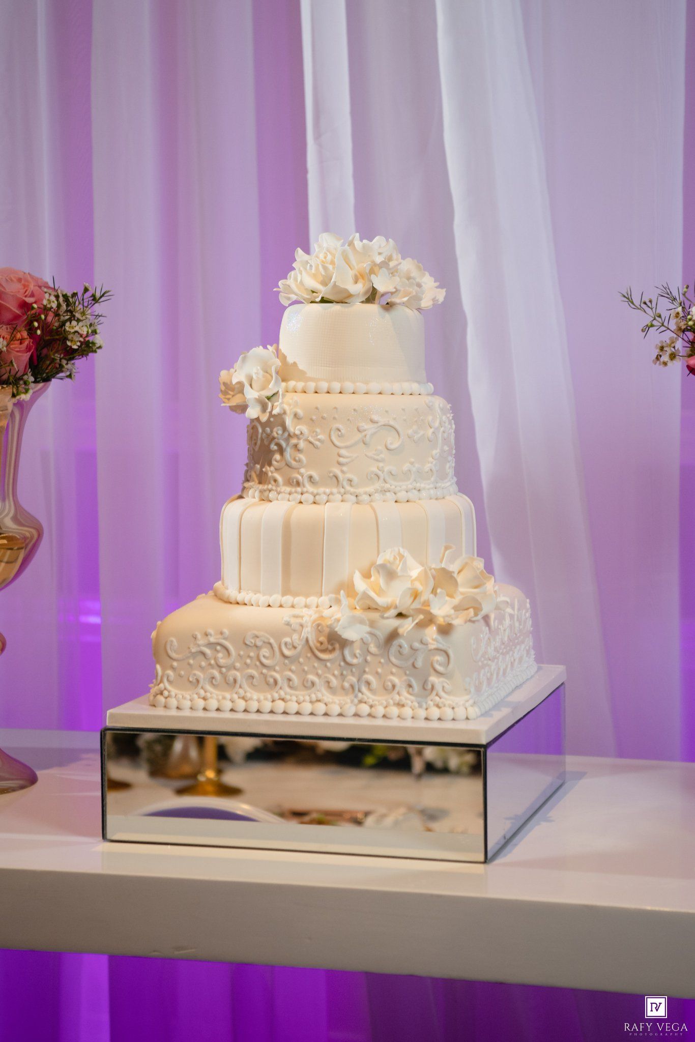 Elegant four-tier white wedding cake with floral decorations on a mirrored stand against purple draped backdrop.