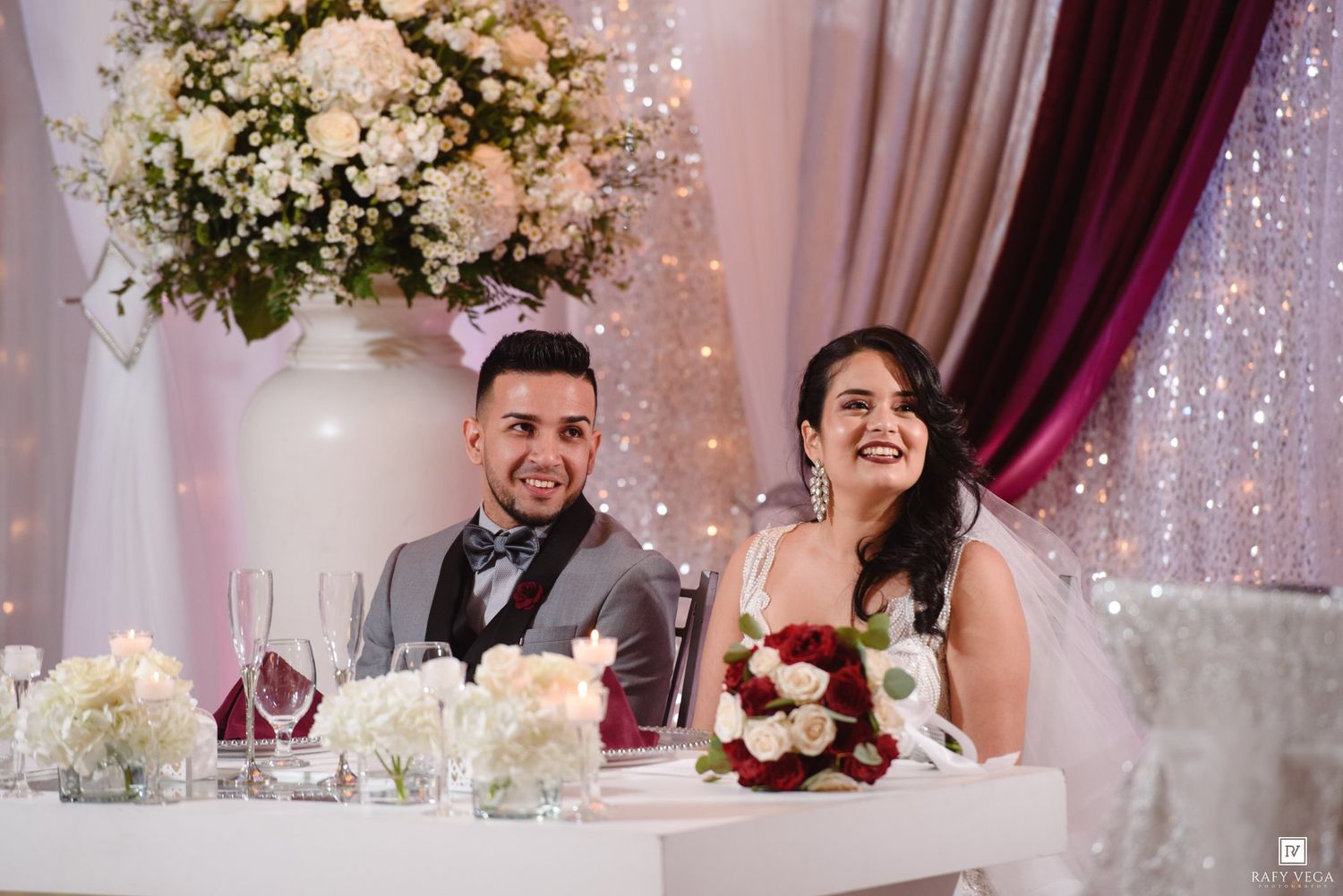 Smiling bride and groom seated at elegant wedding reception table with white floral arrangements and sparkling backdrop.