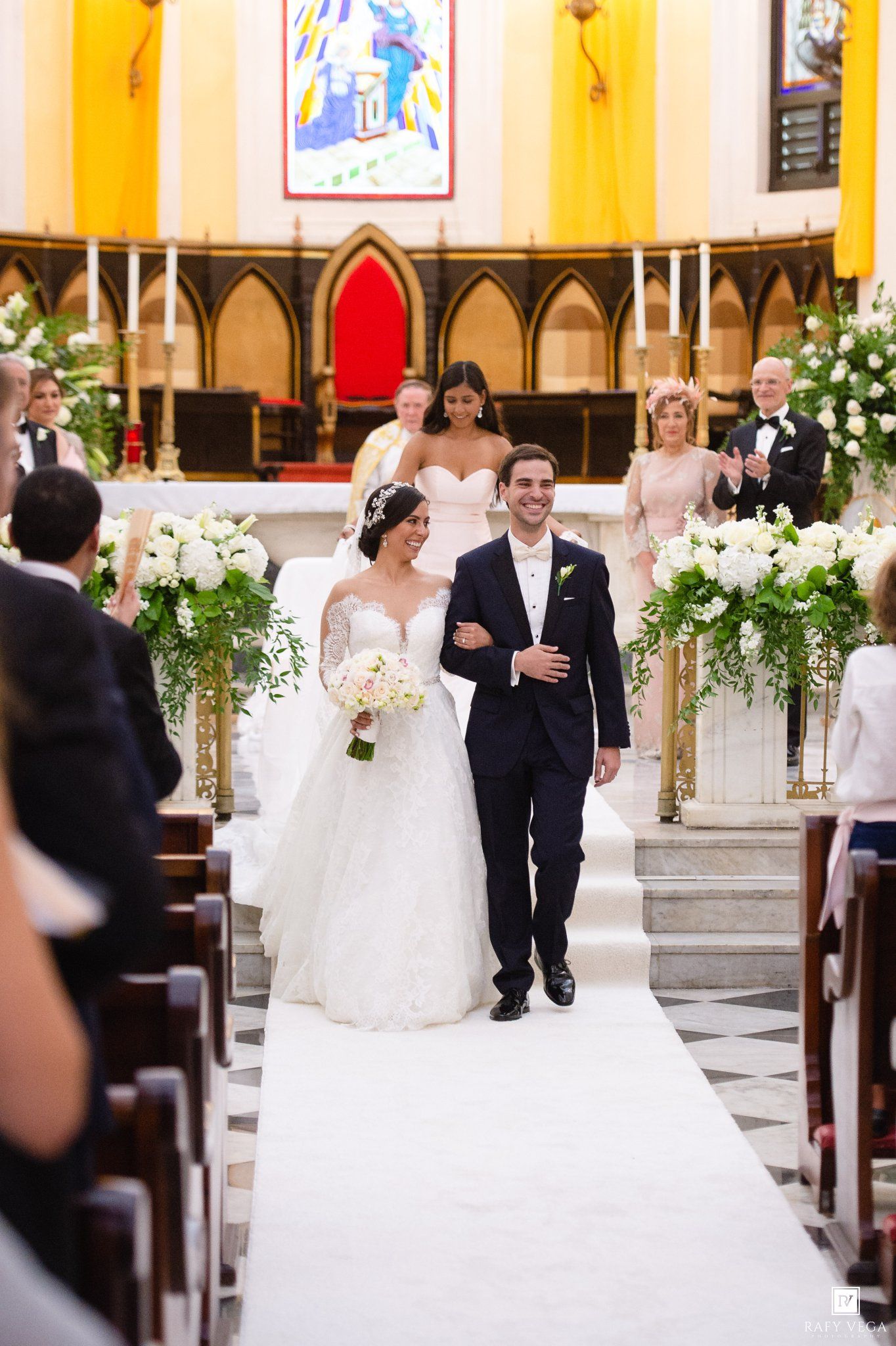 Bride and groom walk down church aisle after wedding ceremony, surrounded by white floral arrangements.
