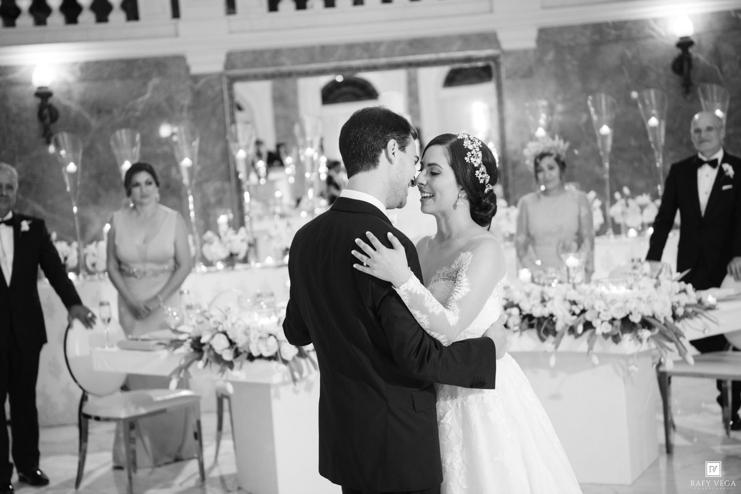 Black and white photo of bride and groom sharing first dance at elegant wedding reception with floral arrangements.