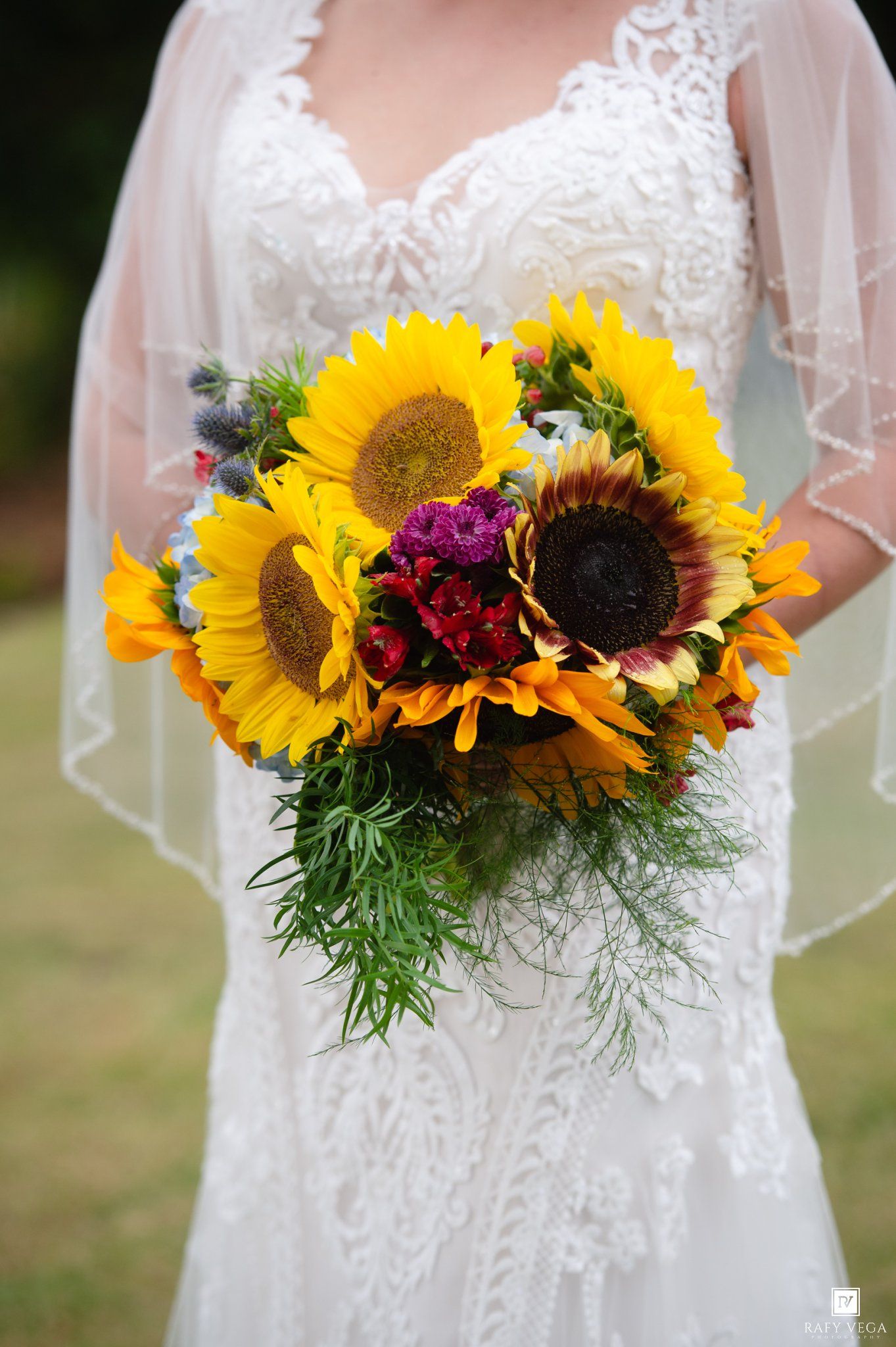 A bride holds a stunning bouquet of bright sunflowers and colorful blooms against her lace wedding gown.