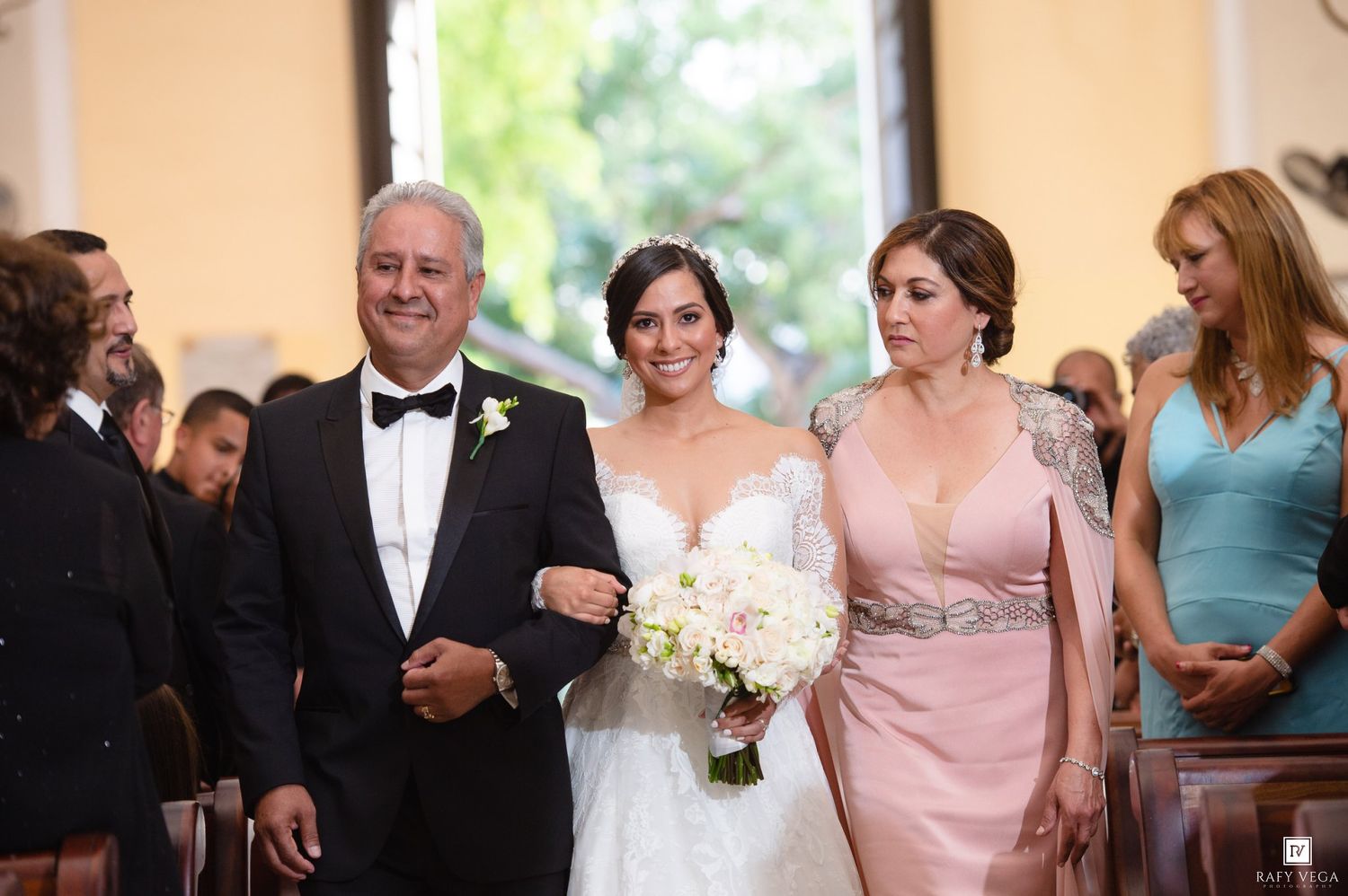 Bride in white lace gown walks down church aisle with father in tuxedo and mother in pink gown.
