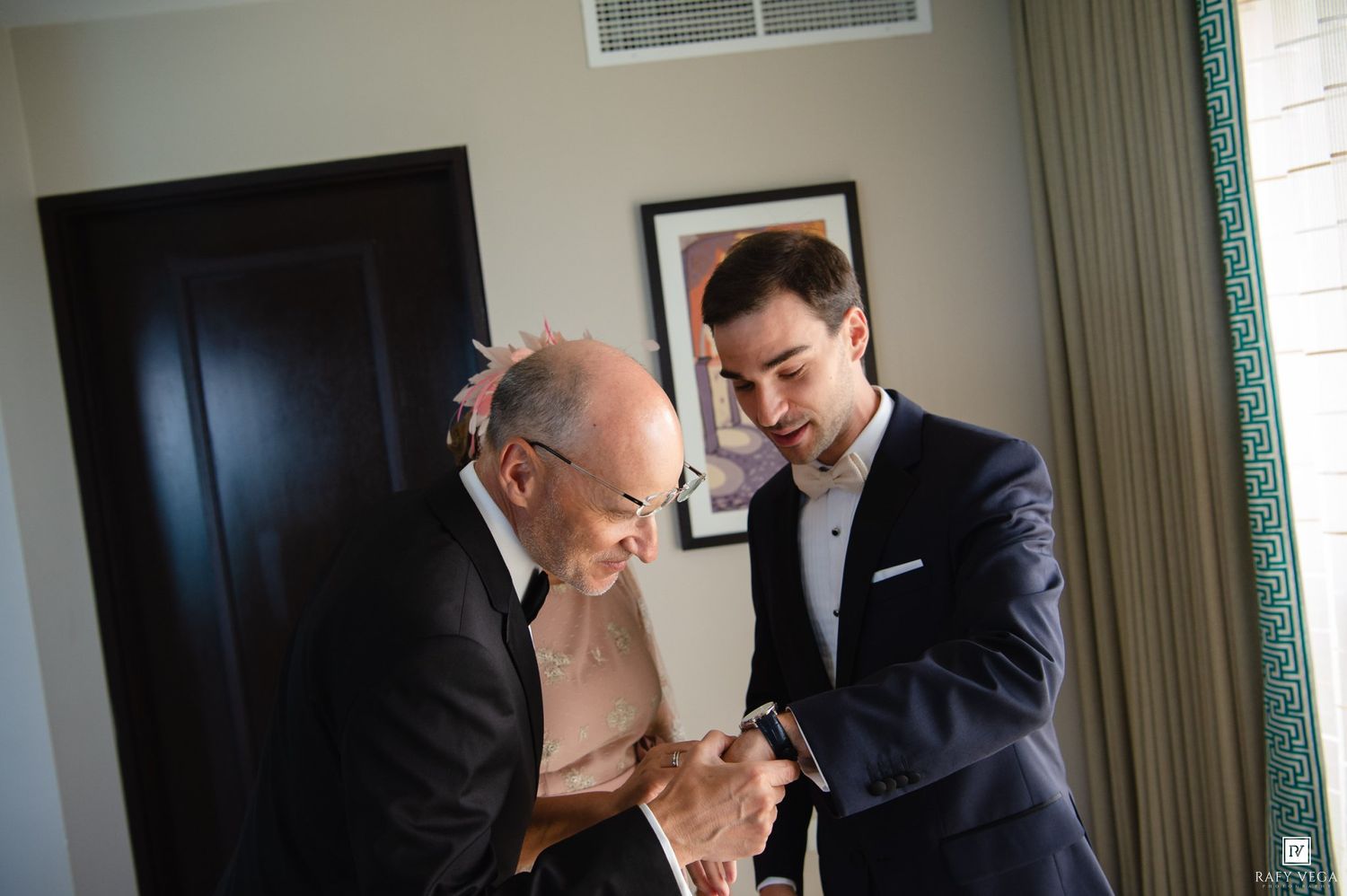 Two men in suits examining something together in an elegant hotel room during a wedding preparation.