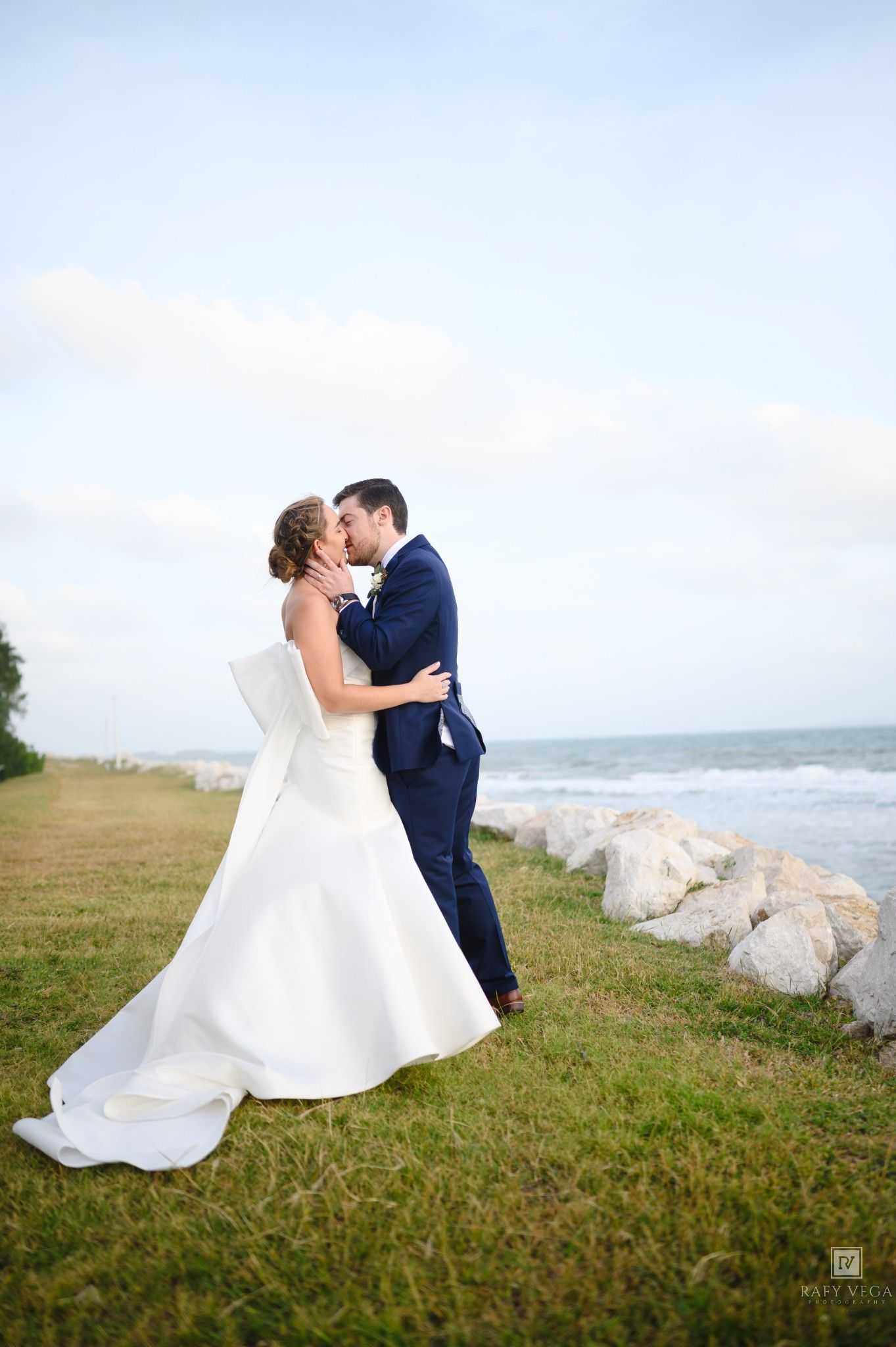 Bride and groom share a romantic kiss by the ocean, wearing a white gown and navy suit on a grassy coastal lawn.