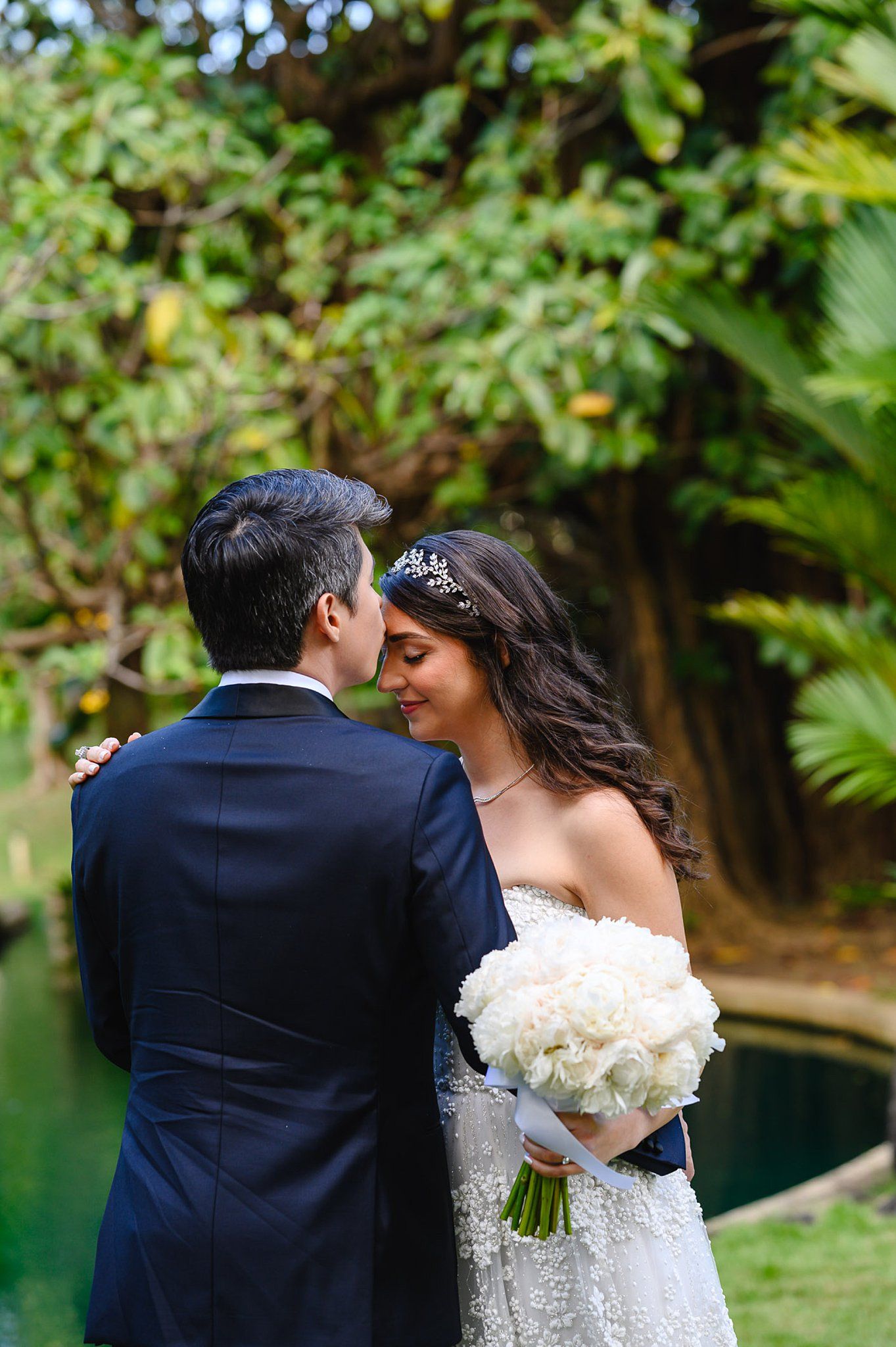 Bride and groom share a romantic moment in a lush garden, bride holding a white floral bouquet wearing a tiara.