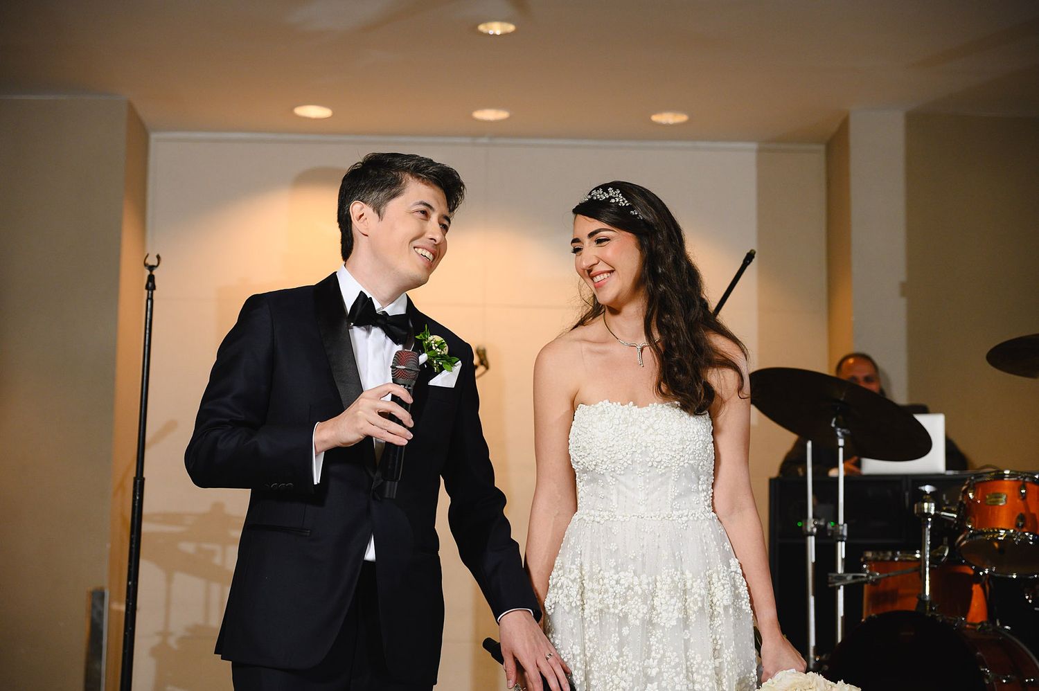 Smiling bride in strapless lace gown and groom in black tuxedo share a joyful moment at their wedding reception.