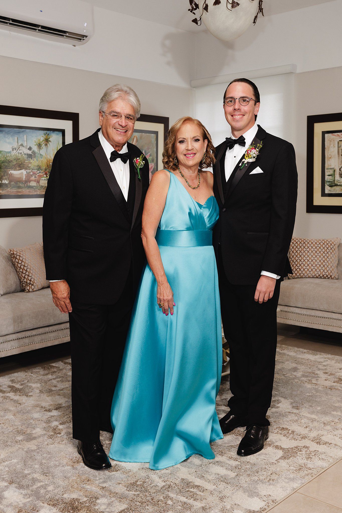 Three elegantly dressed people pose together, two men in tuxedos and a woman in a turquoise gown, at a formal event.