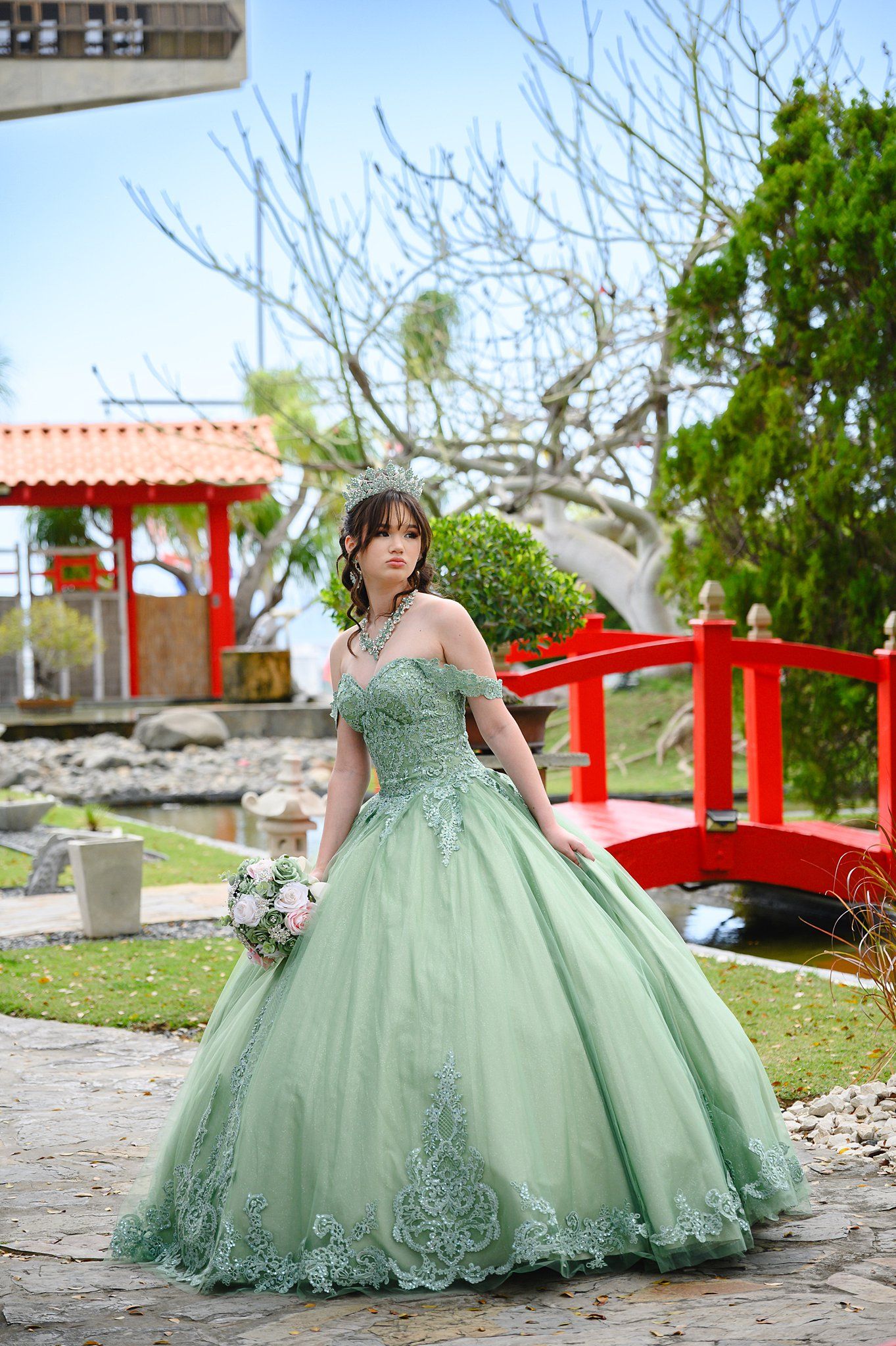 Young woman in sage green off-shoulder ballgown holding bouquet near red Japanese bridge in garden.