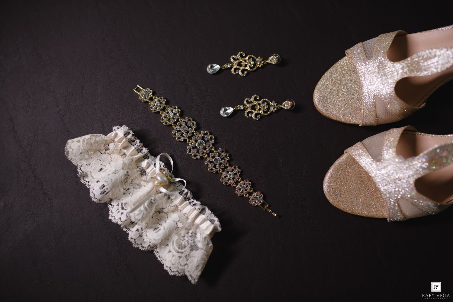 Elegant bridal accessories on dark background featuring lace garter, gold earrings, bracelet, and glittery champagne wedding heels.