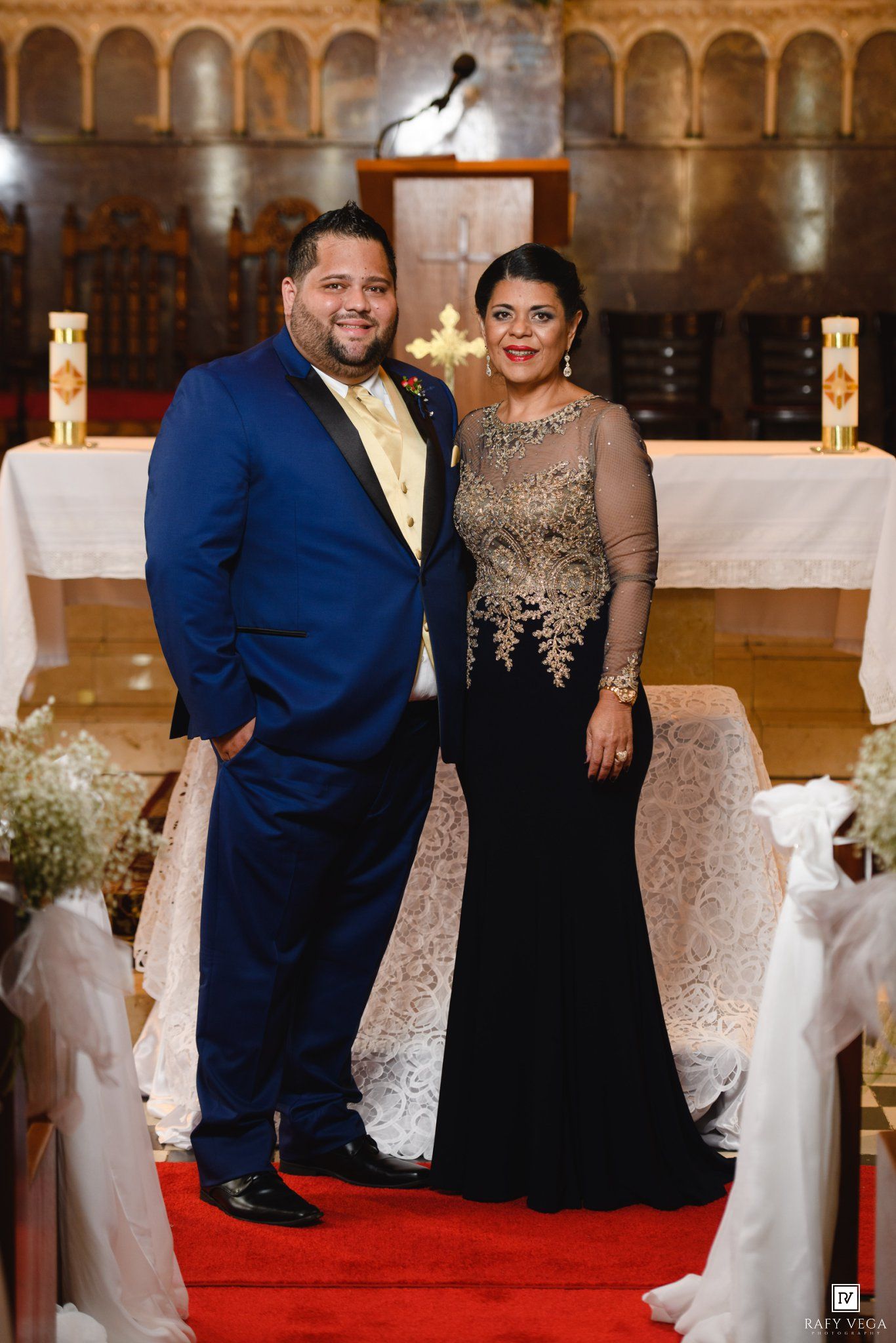 Elegant couple posing in church, man in blue tuxedo, woman in black gold lace gown, red carpet aisle.