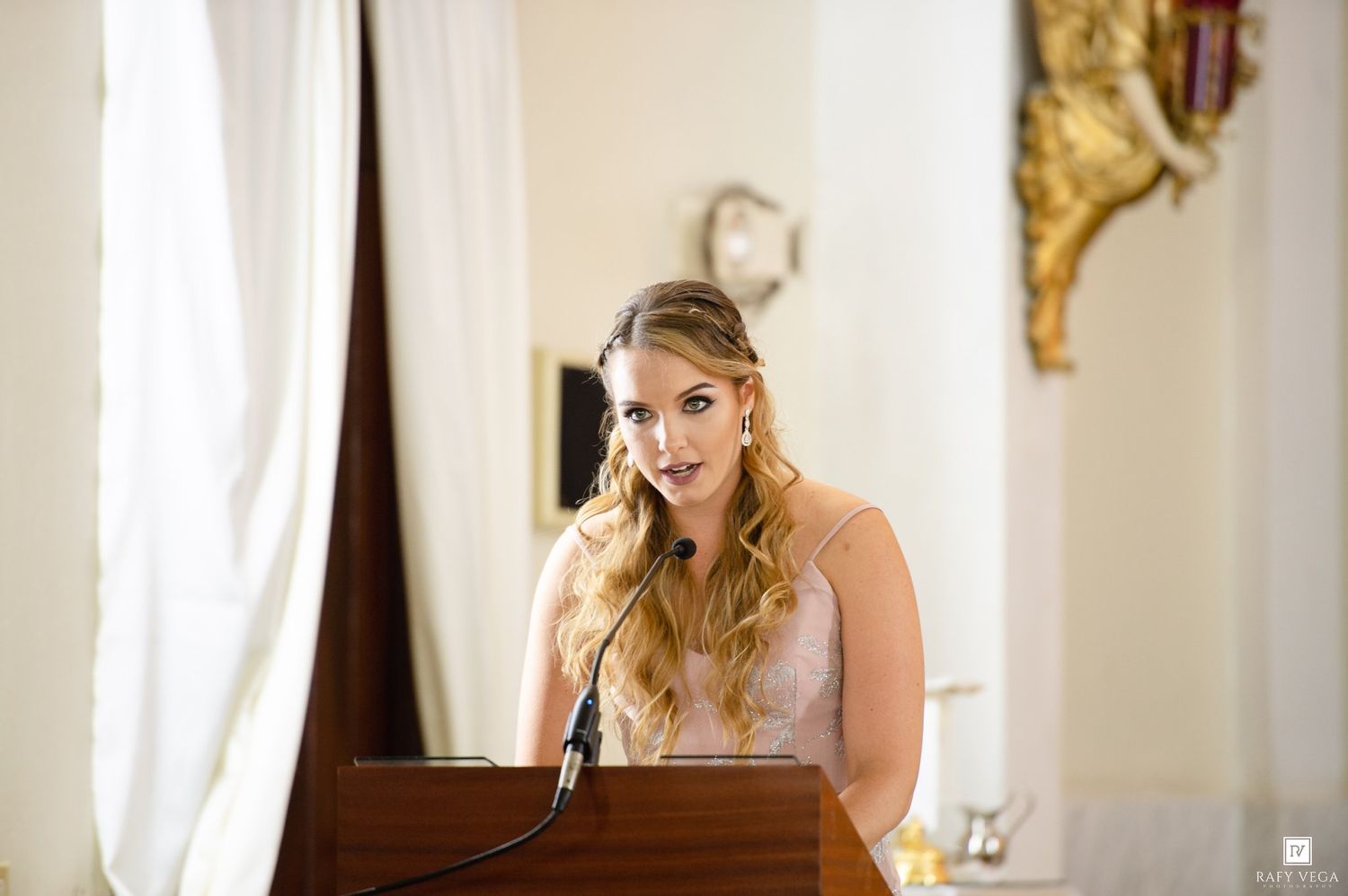 Young woman with blonde wavy hair speaking at a wooden podium in an elegant venue wearing a pink dress.
