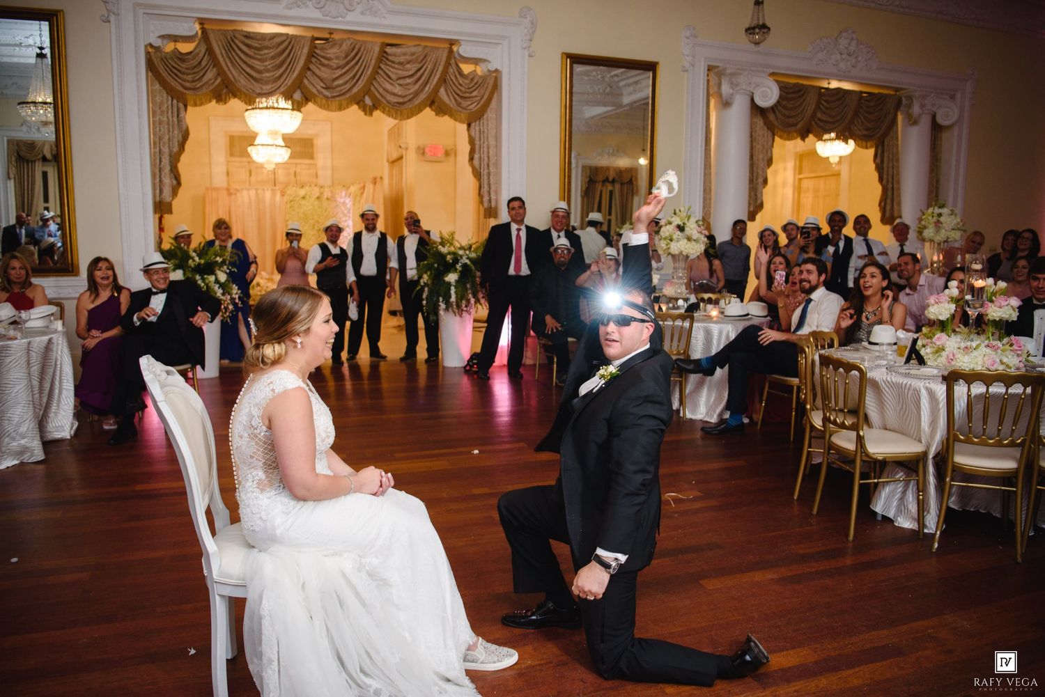 Groom on one knee holds up garter while bride sits in chair at wedding reception.