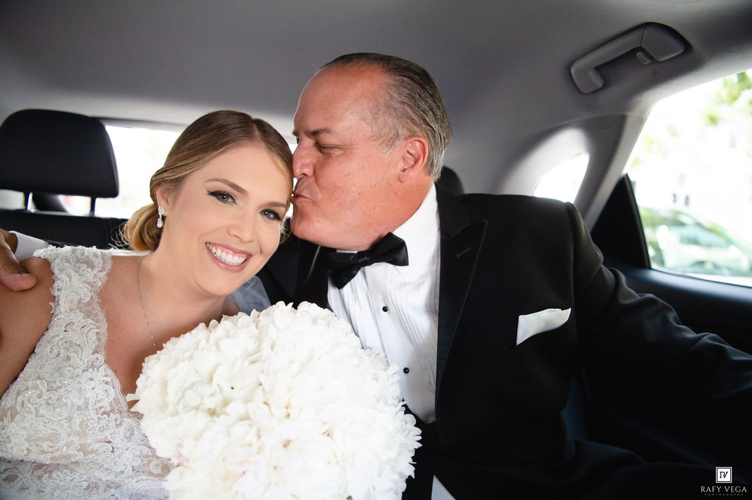 Bride in lace gown holding white bouquet receives kiss on cheek from groom in tuxedo inside a car.