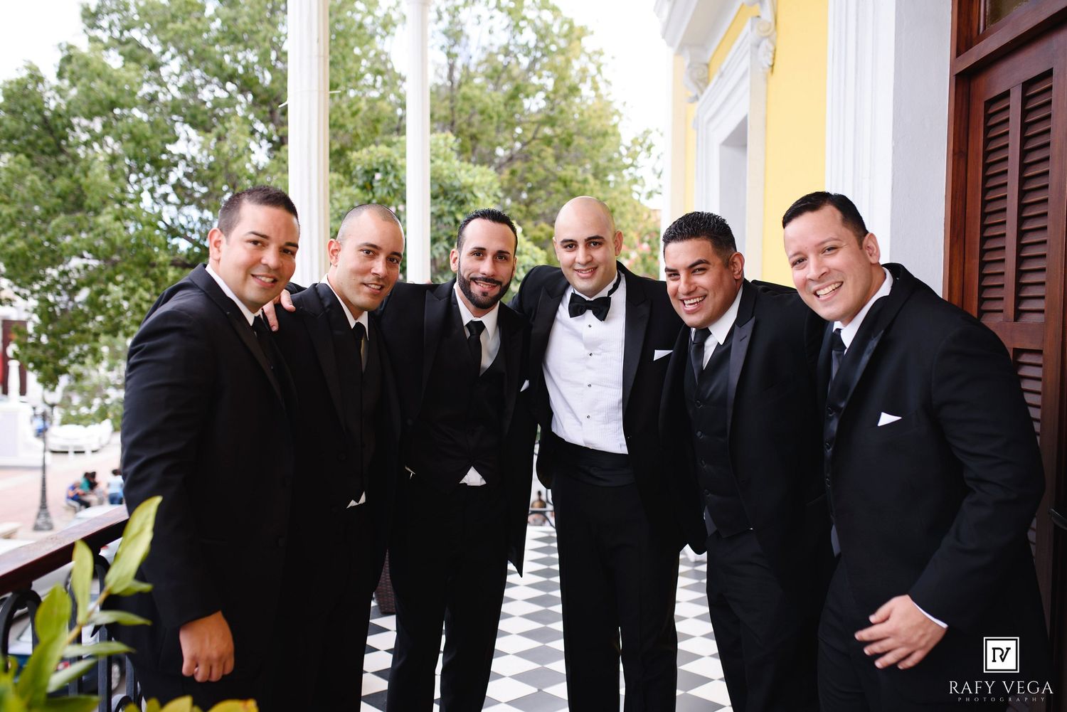 Five groomsmen in black tuxedos smiling together on a decorated outdoor venue balcony at a wedding.