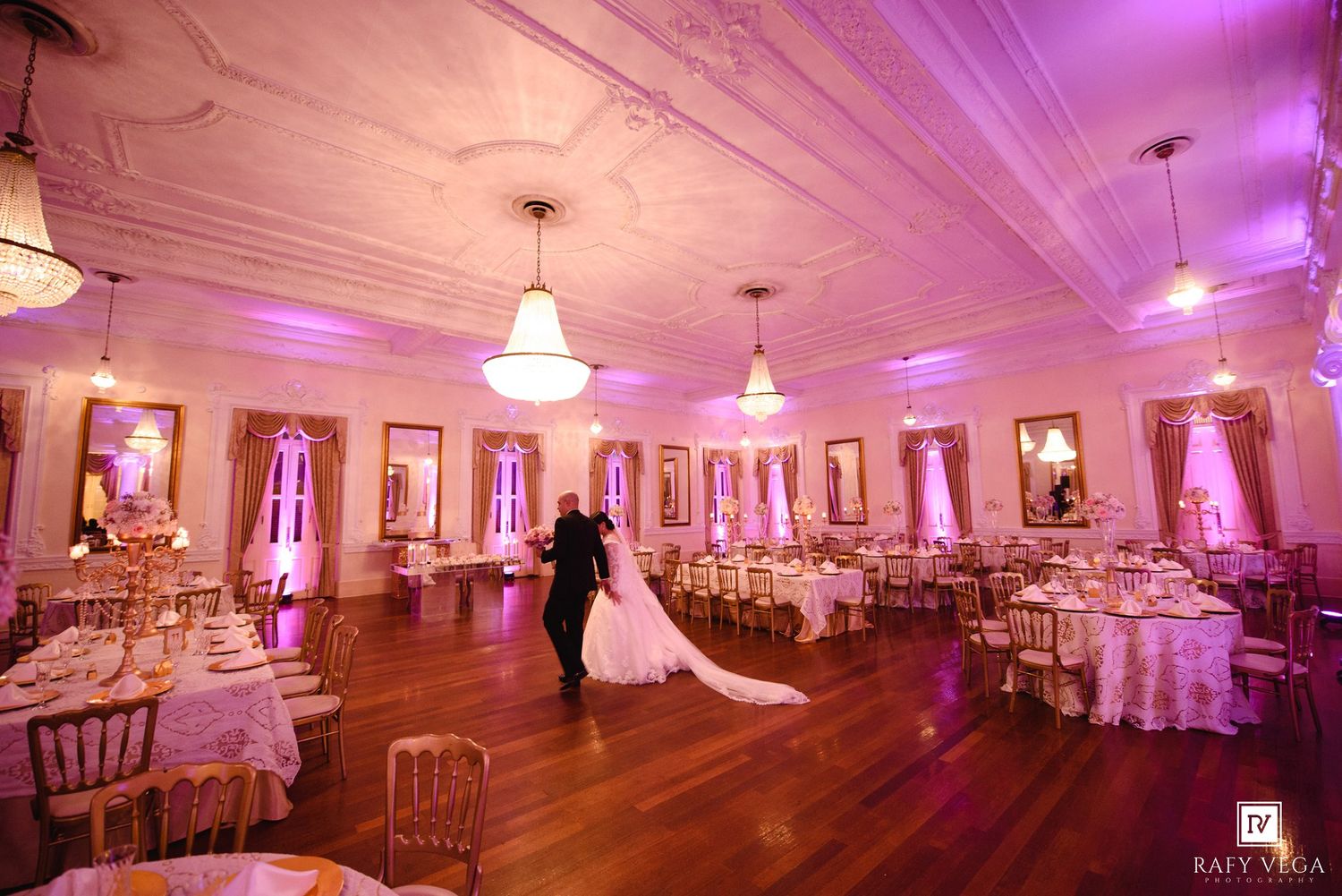Bride and groom share first dance in elegant ballroom with pink uplighting, white tablecloths, and ornate ceiling details.