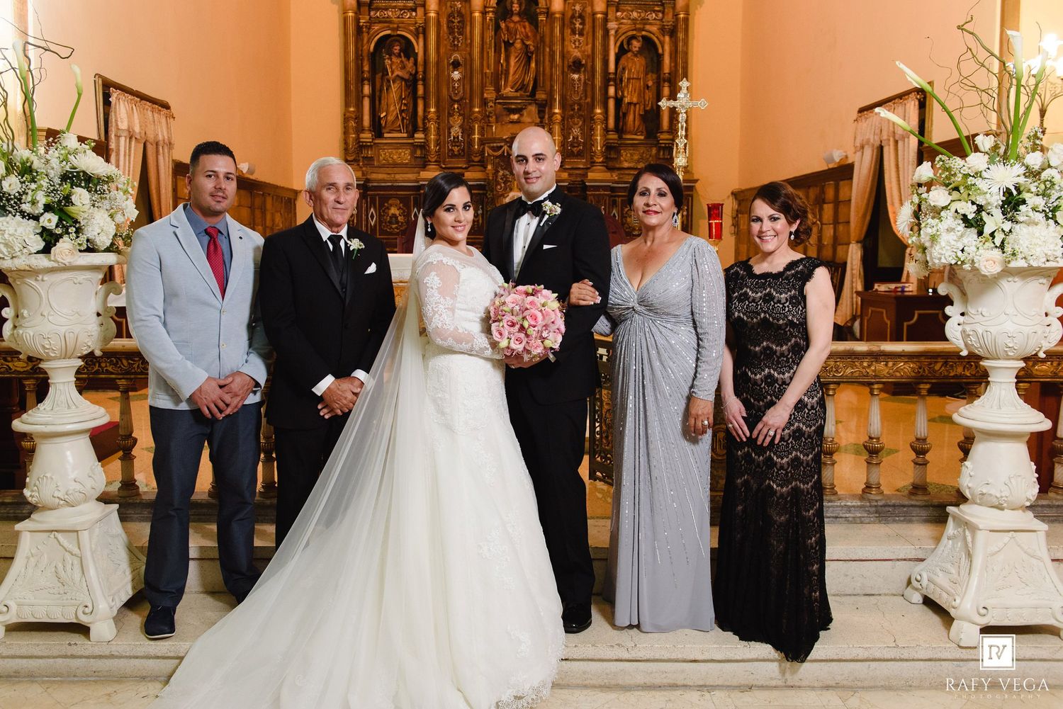 Wedding party poses in elegant church with bride in white gown holding pink bouquet, groom in tuxedo, and four family members.