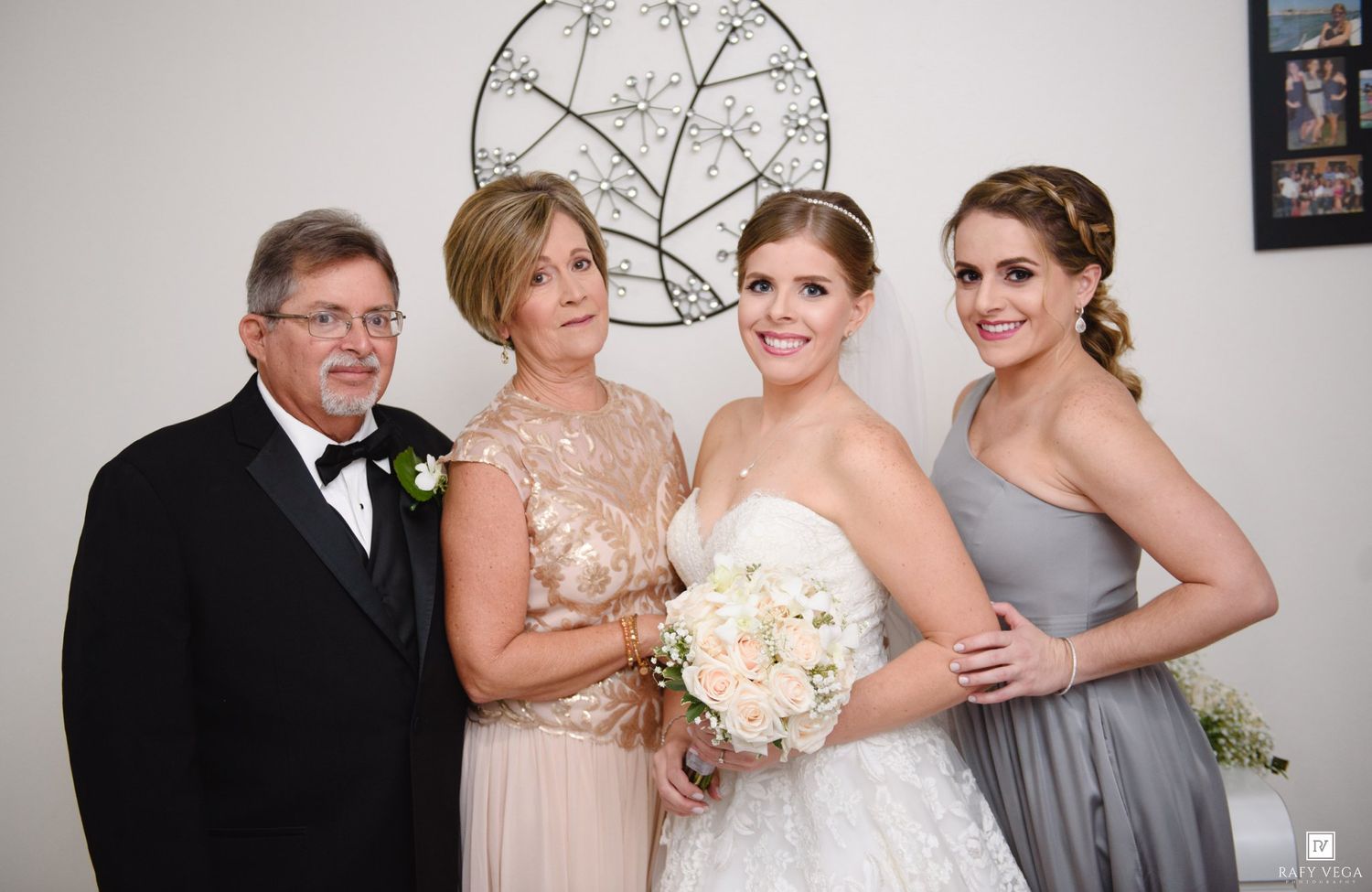 A bride in a white gown holds a bouquet, posing with family including parents and a bridesmaid in gray dress.