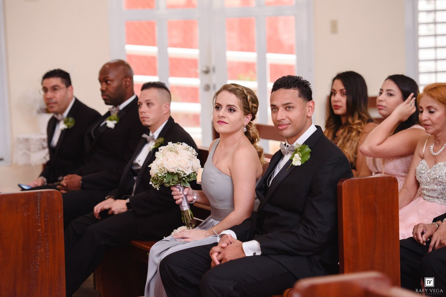 Bride and groom sit together in church pews surrounded by wedding party guests during a formal ceremony.