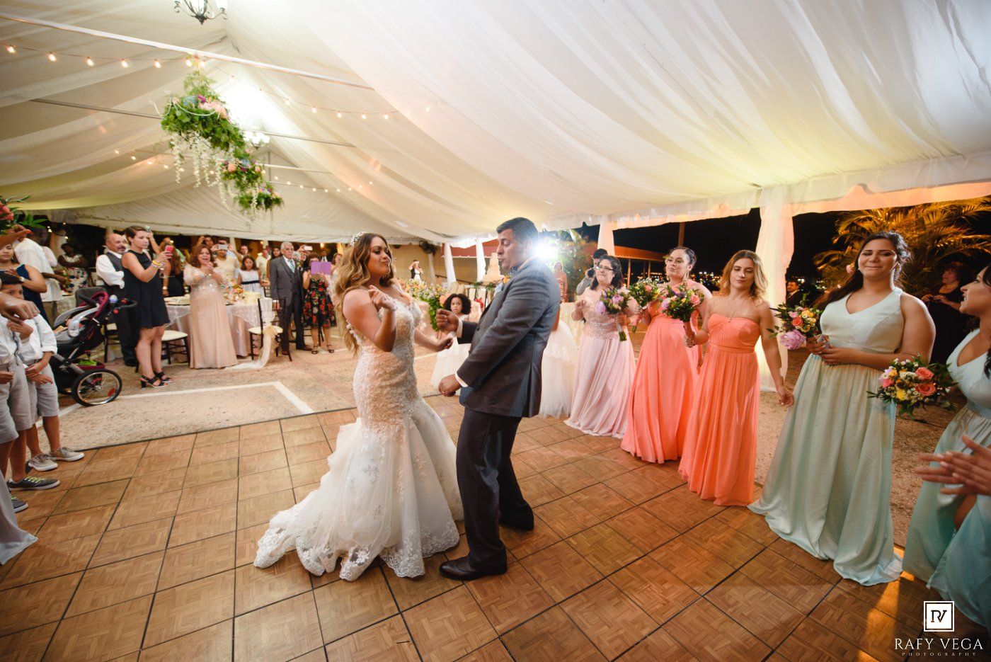 Bride and groom share a joyful first dance surrounded by bridesmaids in coral and mint gowns at an elegant tent reception.