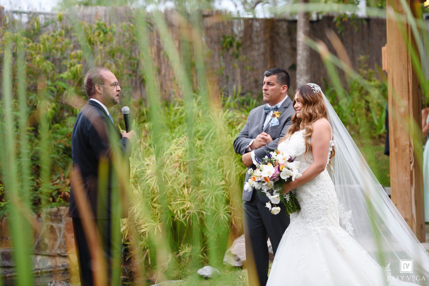 Bride and groom stand with officiant during an outdoor garden wedding ceremony surrounded by lush greenery.