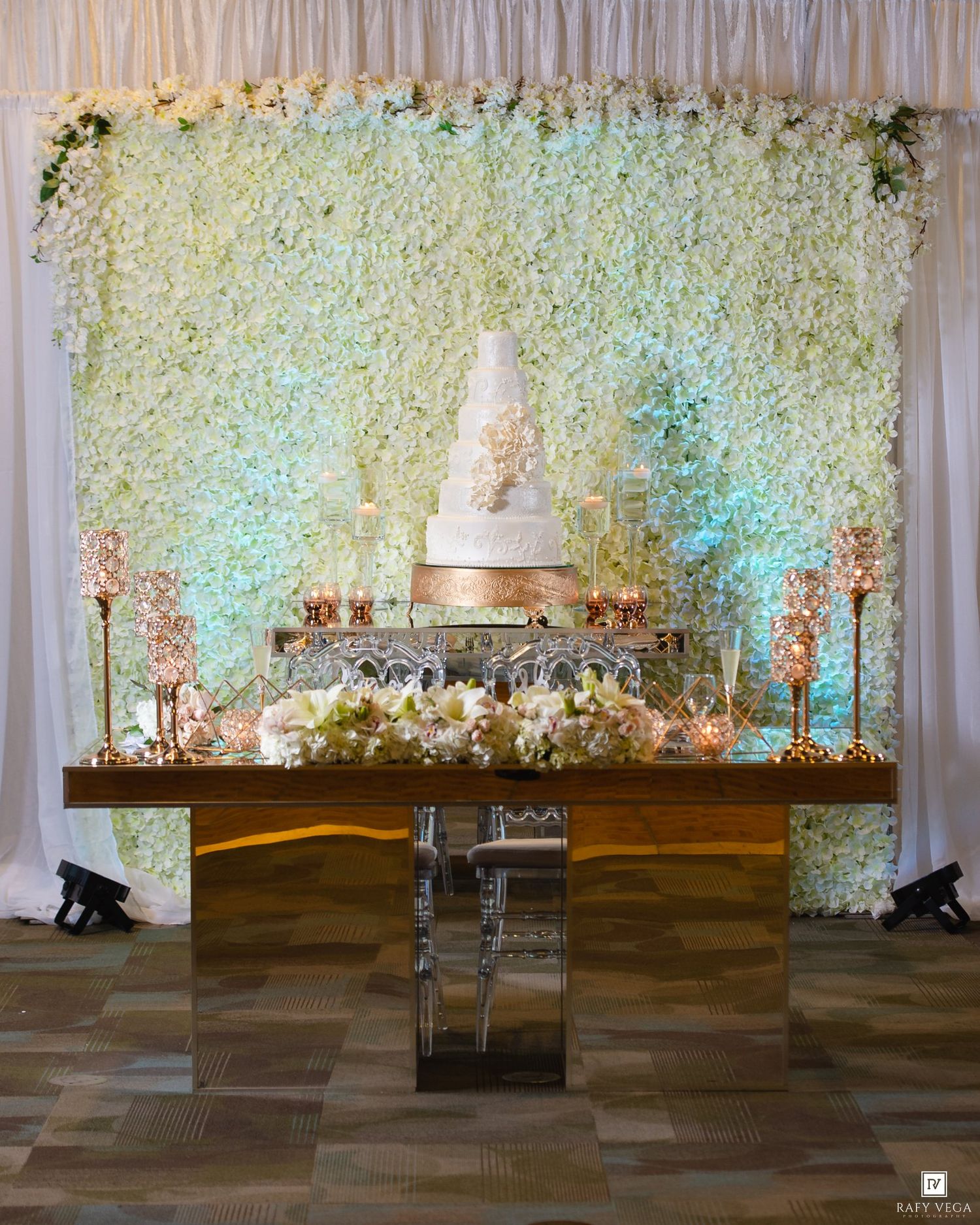 Elegant wedding cake display on mirrored table with white floral wall backdrop and gold candelabras.